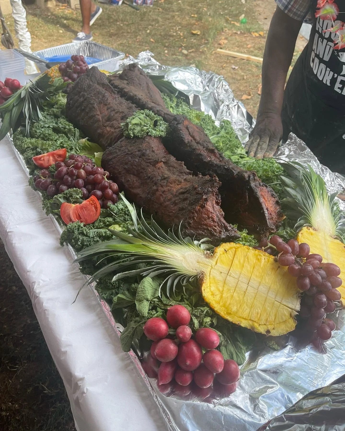 Barbecue ribs on a tray decorated with pineapple slices, grapes, cherry tomatoes, and leafy greens, with aluminum foil underneath.