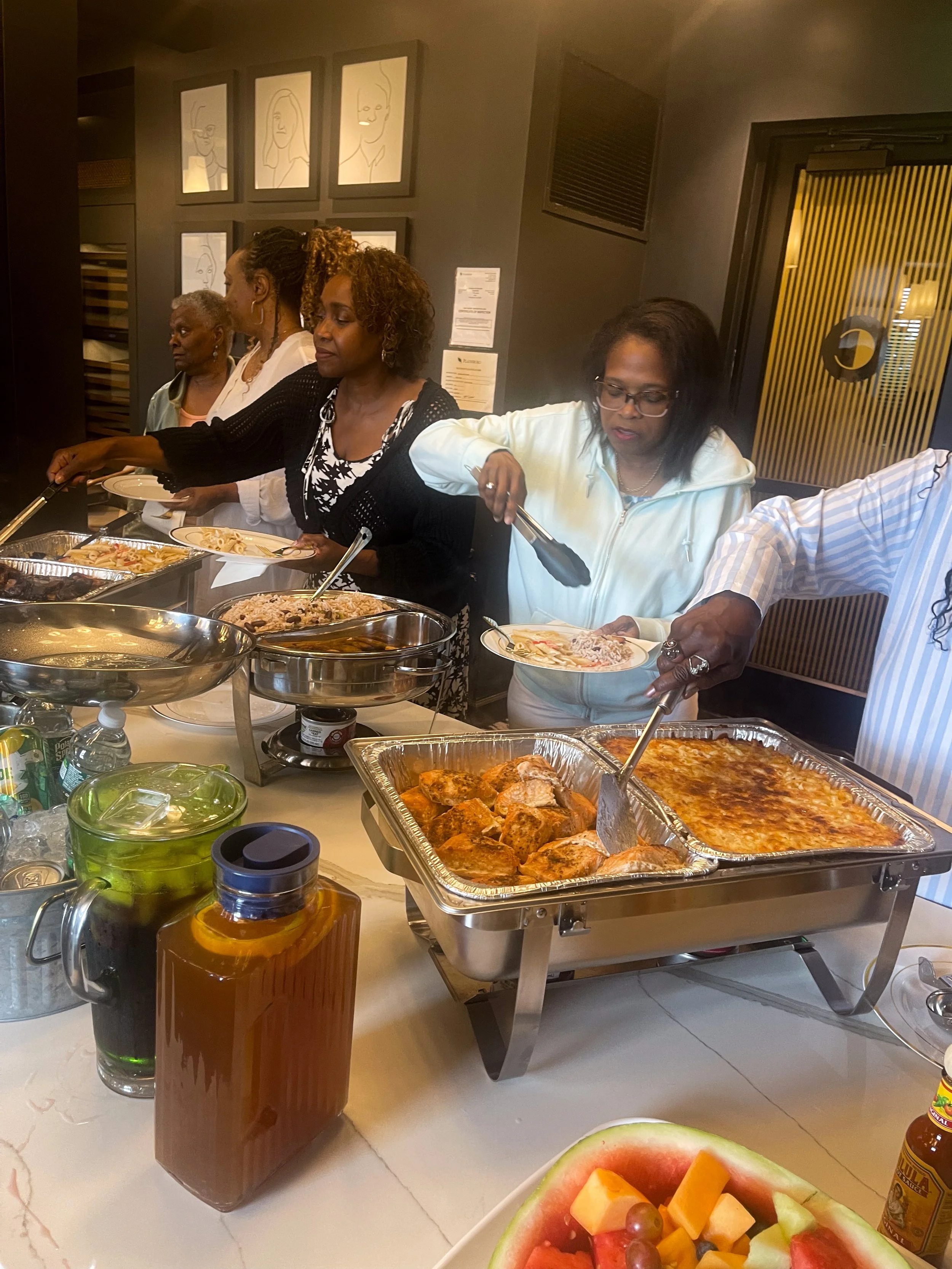 Four women serving themselves food from a buffet table in a restaurant or banquet setting. The table has chafing dishes with various hot foods, and there is a fruit platter with watermelon, cantaloupe, grapes, and other sliced fruits in the foreground.