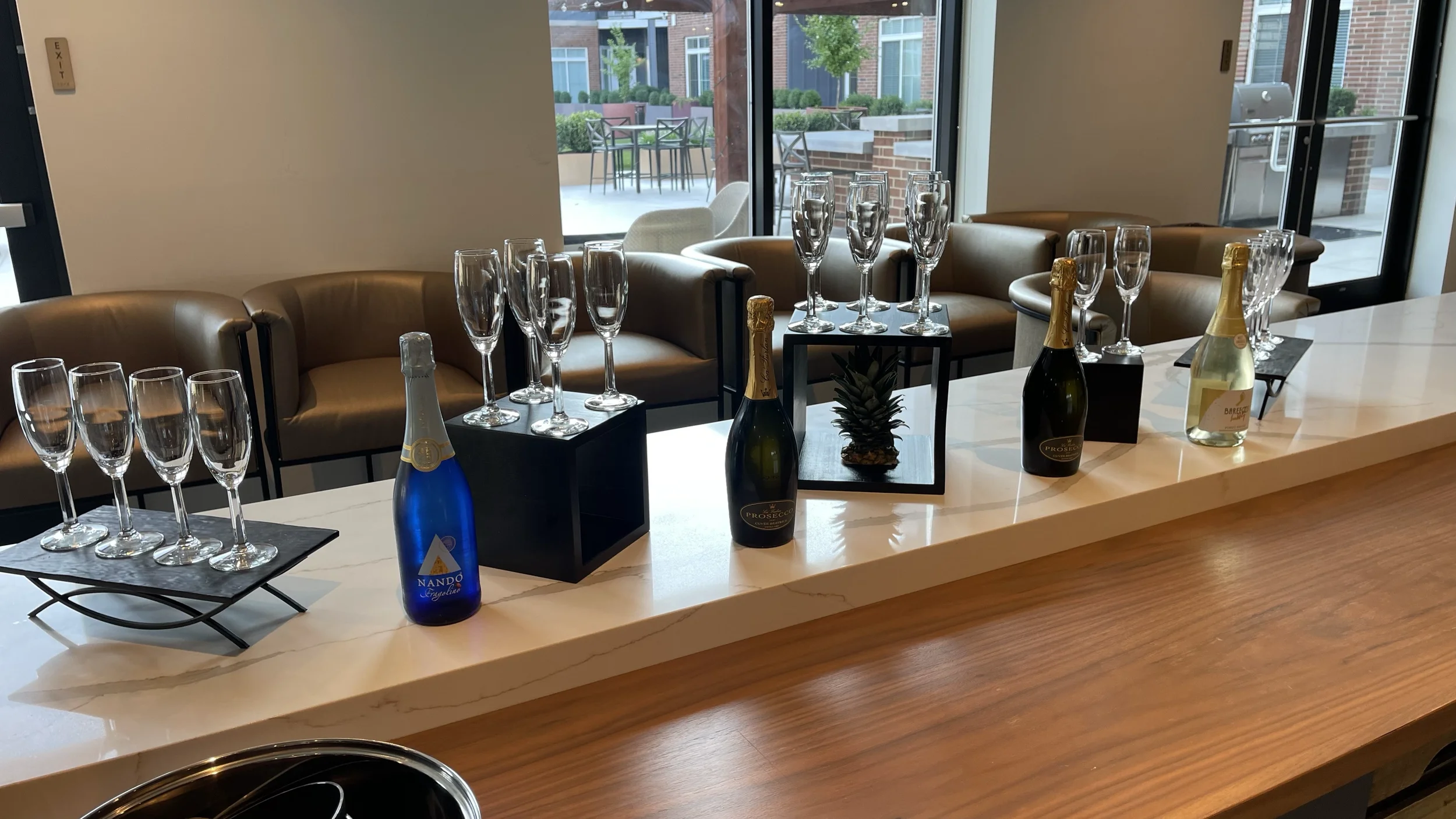Counter with three bottles of sparkling wine, empty champagne glasses, and decorative pineapple, with chairs and large windows in the background.
