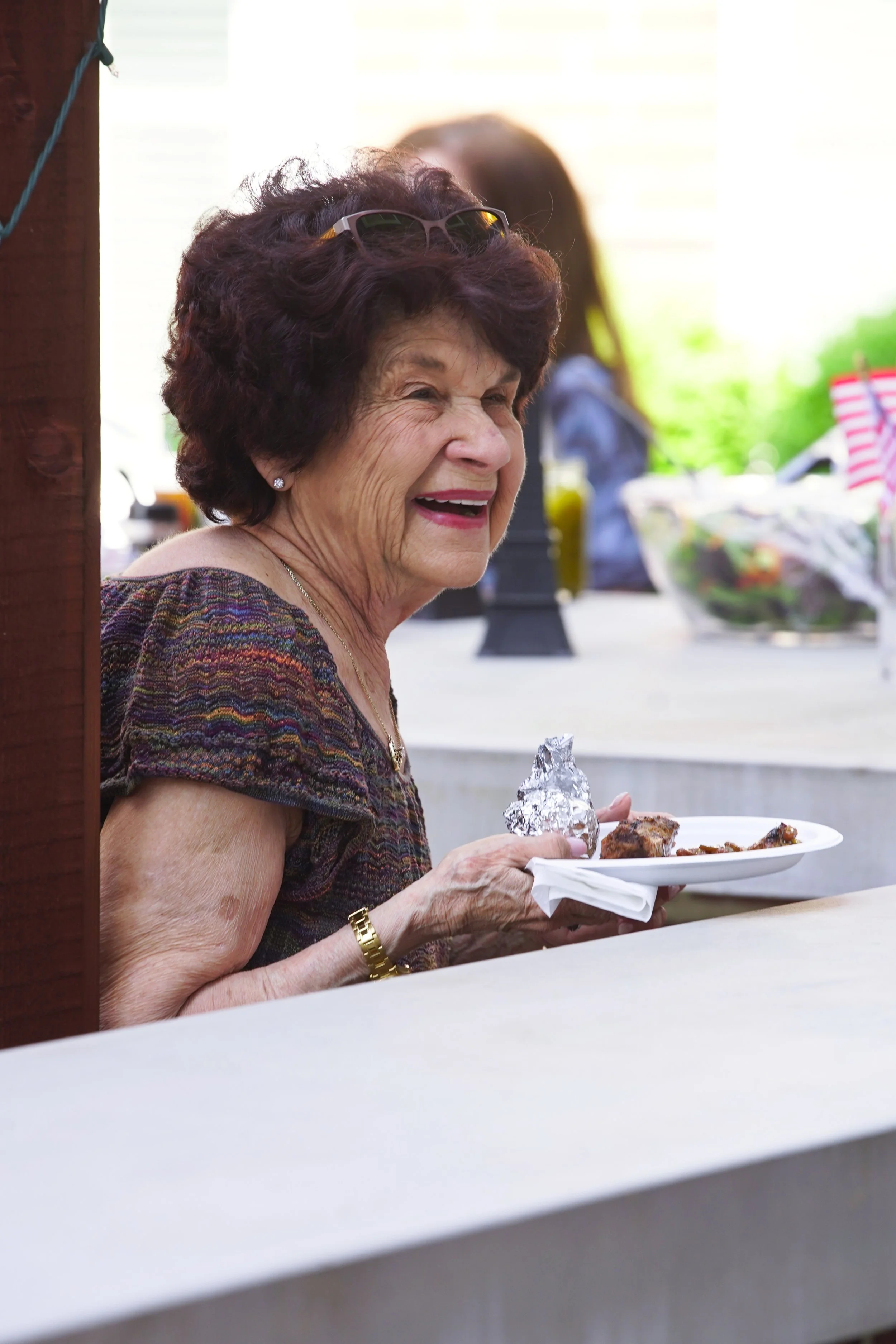 An elderly woman with short curly hair and sunglasses perched on her head is laughing and holding a plate of food, including a piece of foil-wrapped item, at an outdoor gathering.