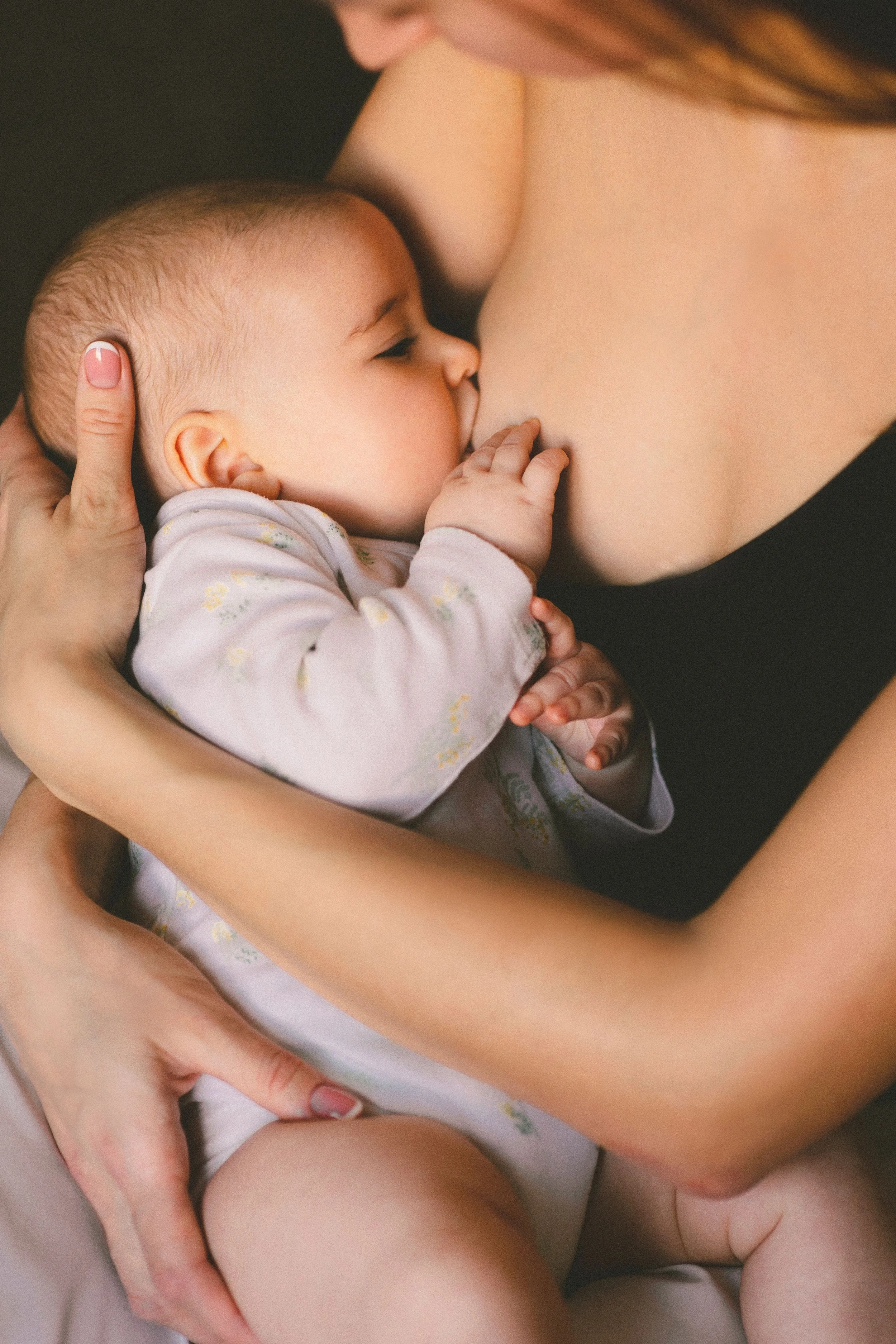 A baby with closed eyes being breastfed by an adult, with the adult's arm and hand supporting the baby.