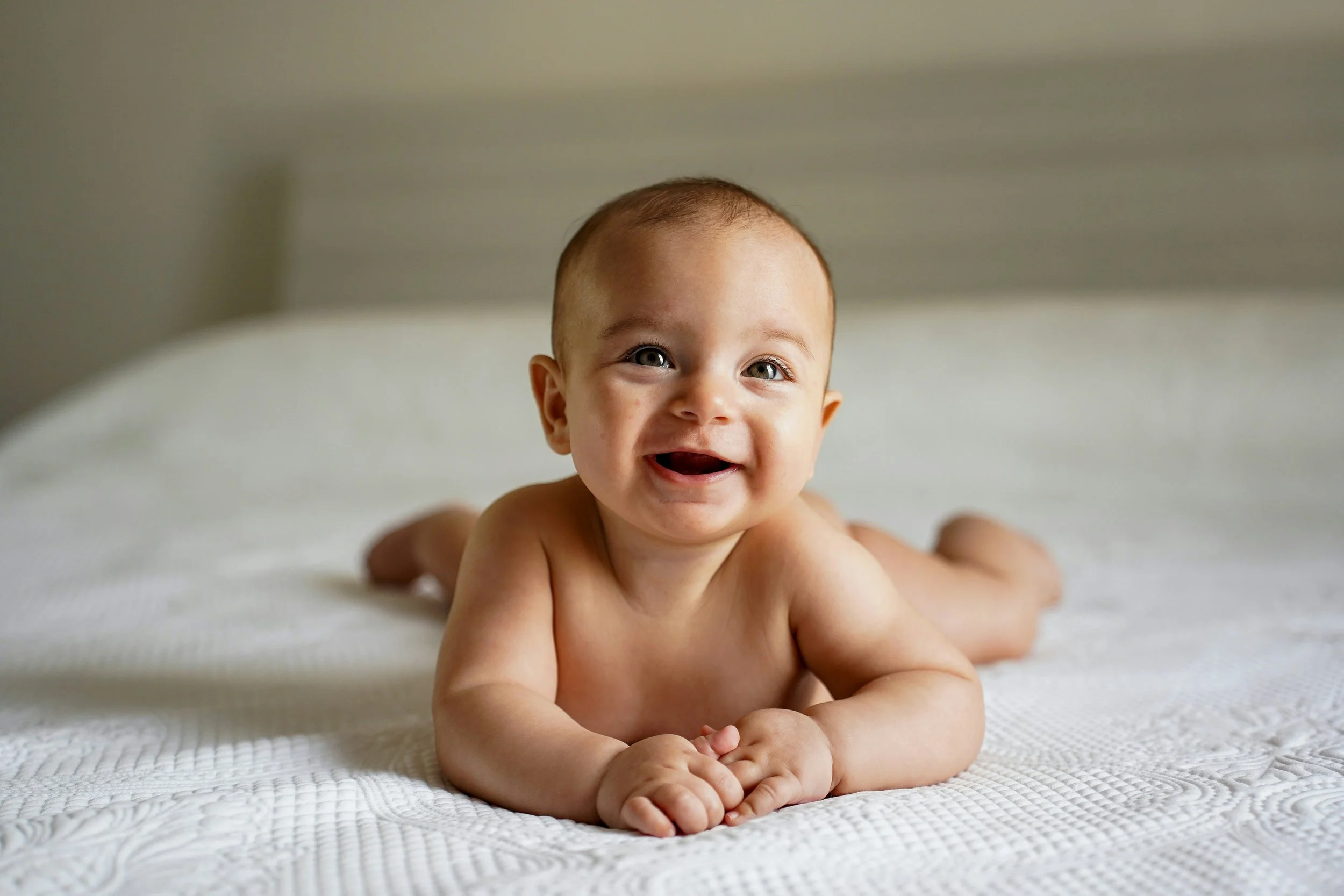 A smiling baby with light skin, lying on a textured white blanket, with short light brown hair, wide eyes, and an open mouth showing two small teeth.