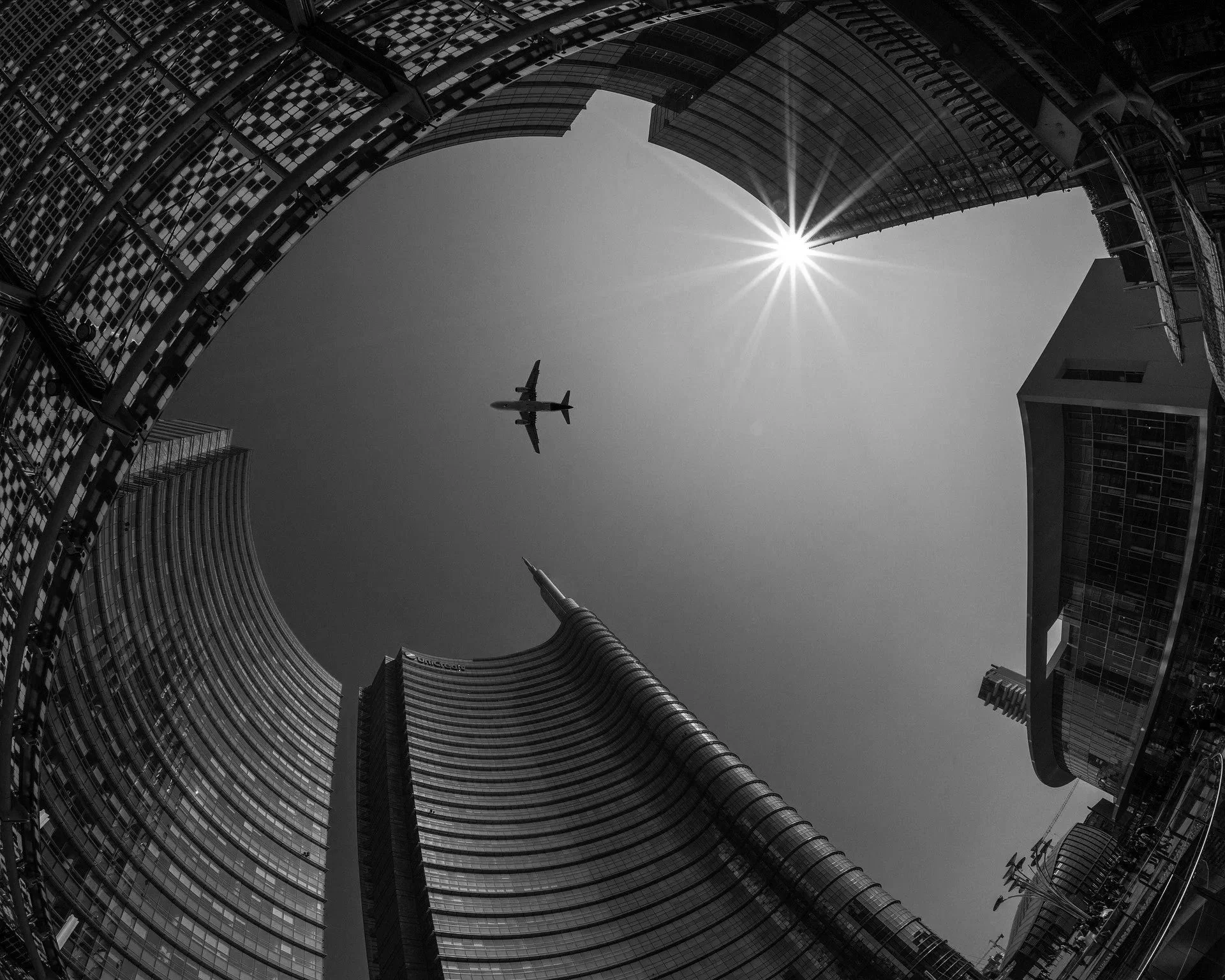 Looking up at modern skyscrapers with glass facades in a city, with the sun shining through, and an airplane flying overhead.