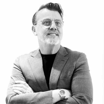 A man with glasses, a beard, and styled hair, wearing a blazer and a watch, smiling with crossed arms, in black and white.