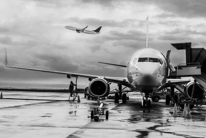 A commercial airplane parked at an airport gate with a jet bridge connected, and a smaller airplane flying in the cloudy sky above.