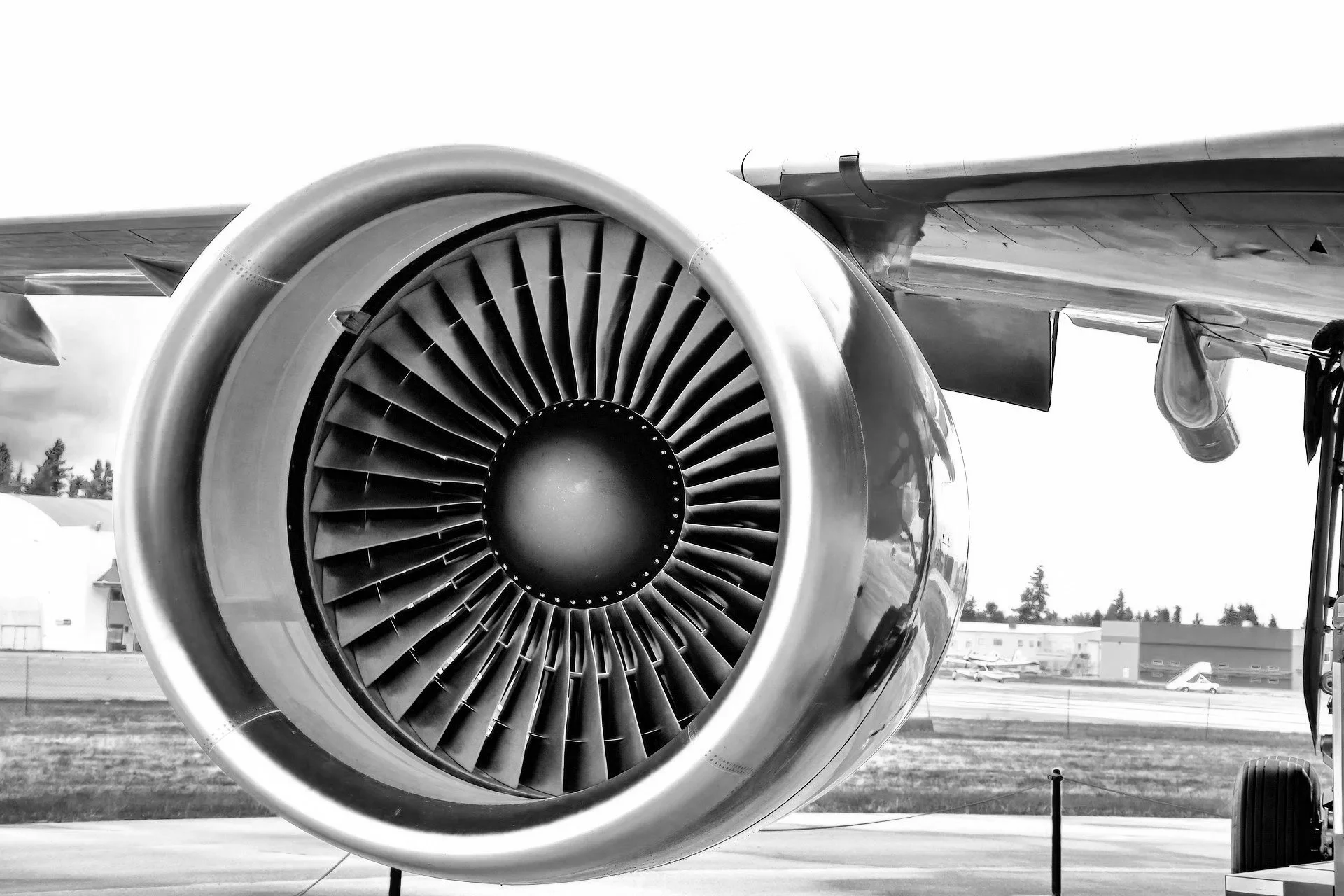 Black and white photo of an airplane jet engine mounted on the wing, with the airplane on the tarmac at an airport.