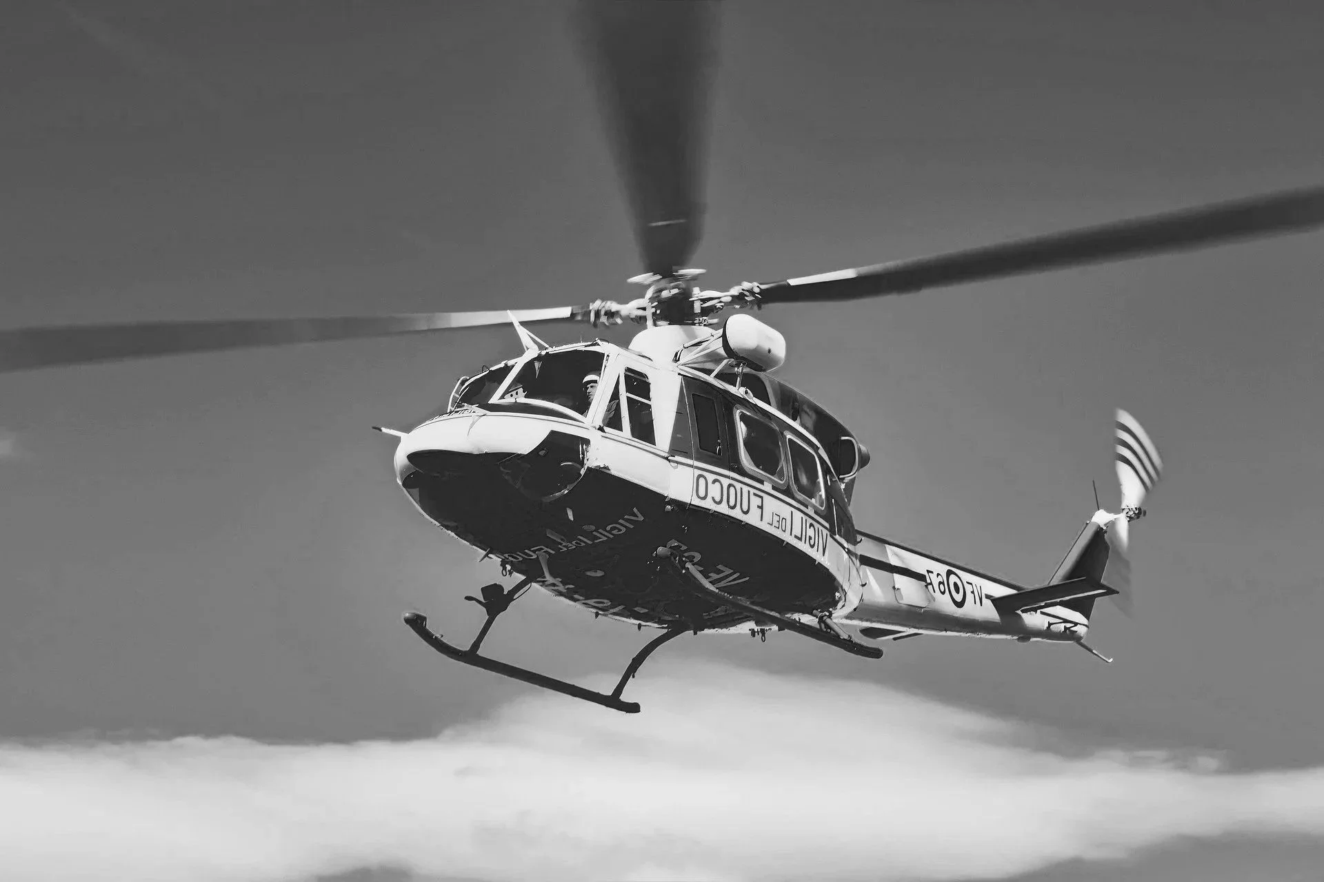 Black and white photo of a helicopter flying in the sky, viewed from below, with visible rotor blades and tail, and a person inside the cockpit.