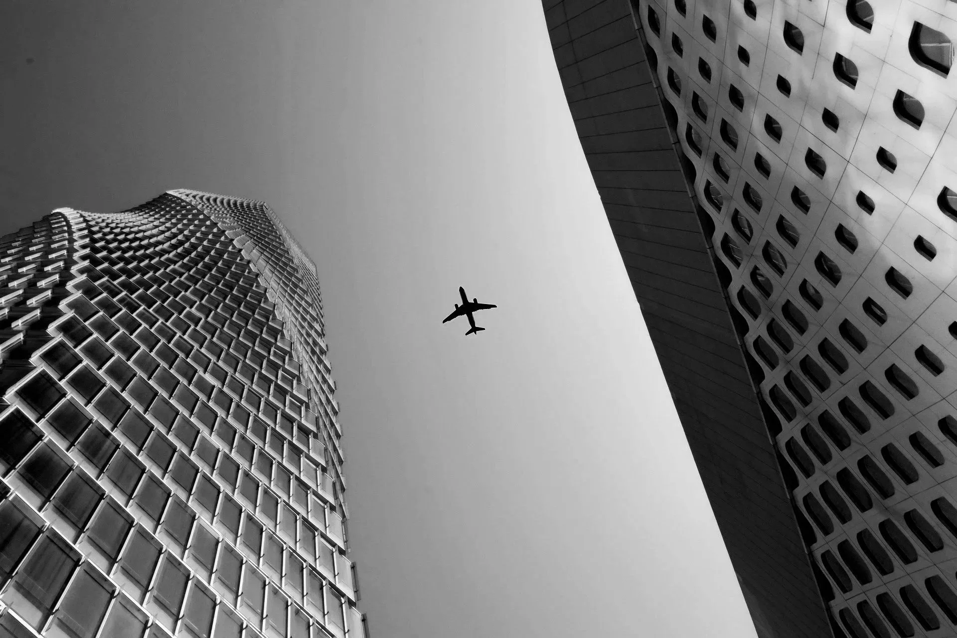Black and white photo of two tall modern buildings with distinctive patterns, viewed from below, with an airplane flying overhead in the sky between them.