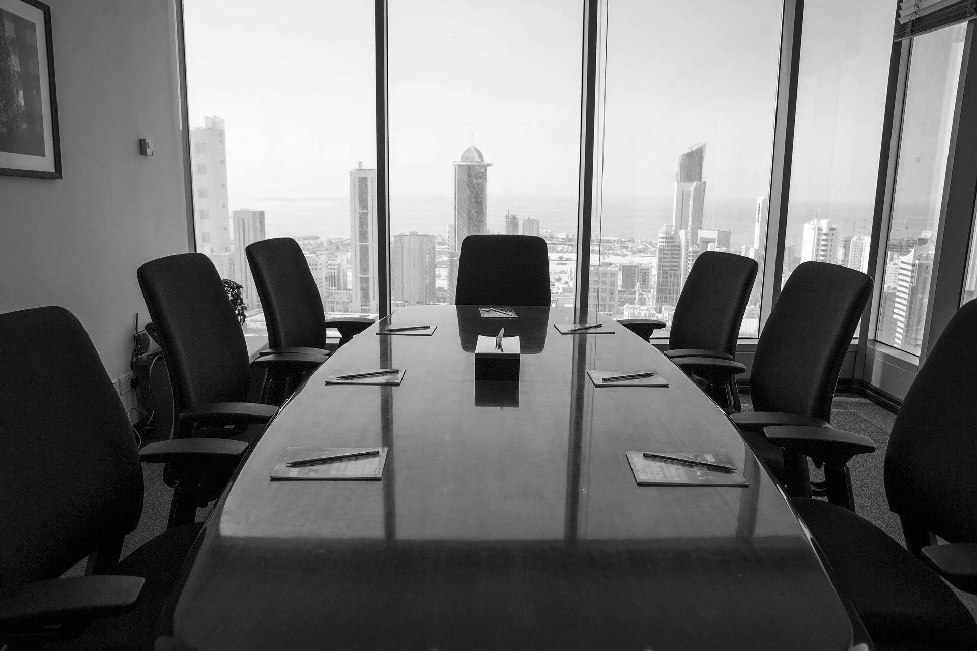 A conference room with a long table and eight chairs, set with notepads and pens, and large windows showing a cityscape skyline.