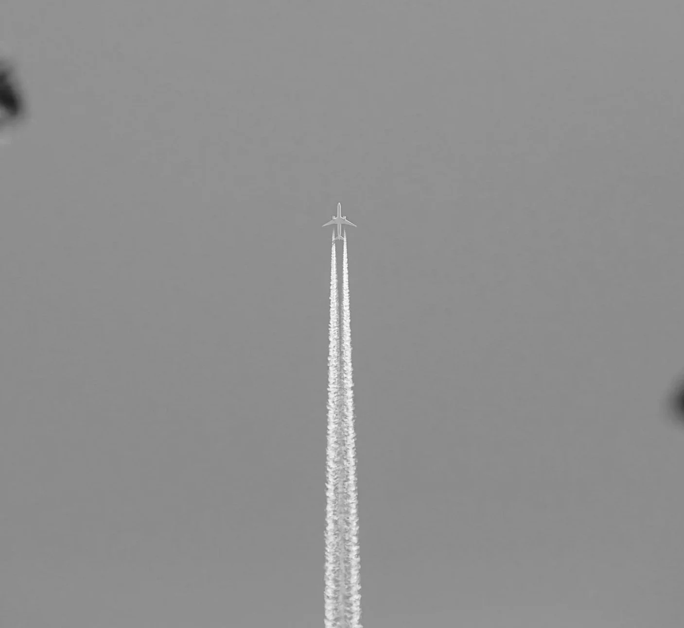 An airplane flying high in the sky leaving contrails behind, with blurred cloud edges visible at the corners.