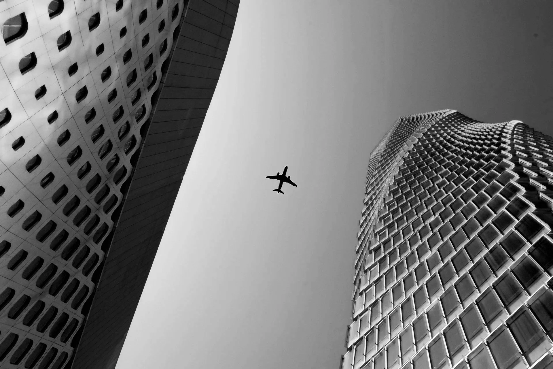 Black and white photo of tall modern buildings with an airplane flying above.