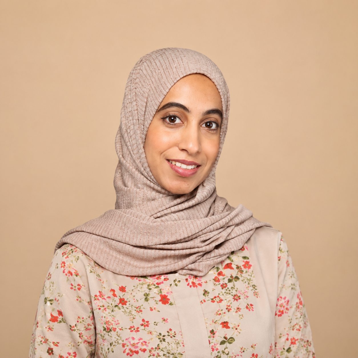 Young woman wearing a beige hijab and floral blouse smiling against a neutral background.