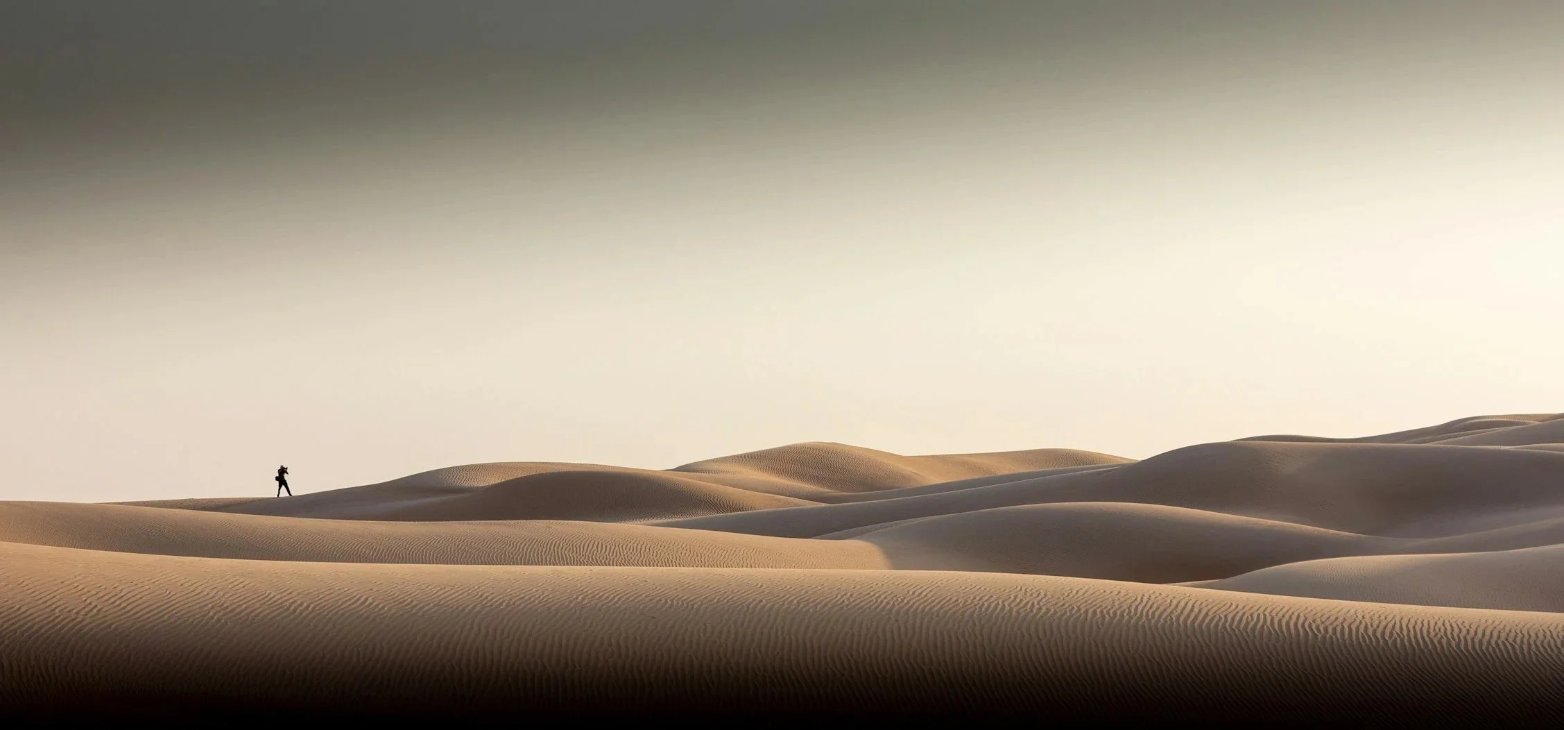 A person walking alone across sand dunes in a desert landscape during sunset or sunrise, with a clear sky in the background.
