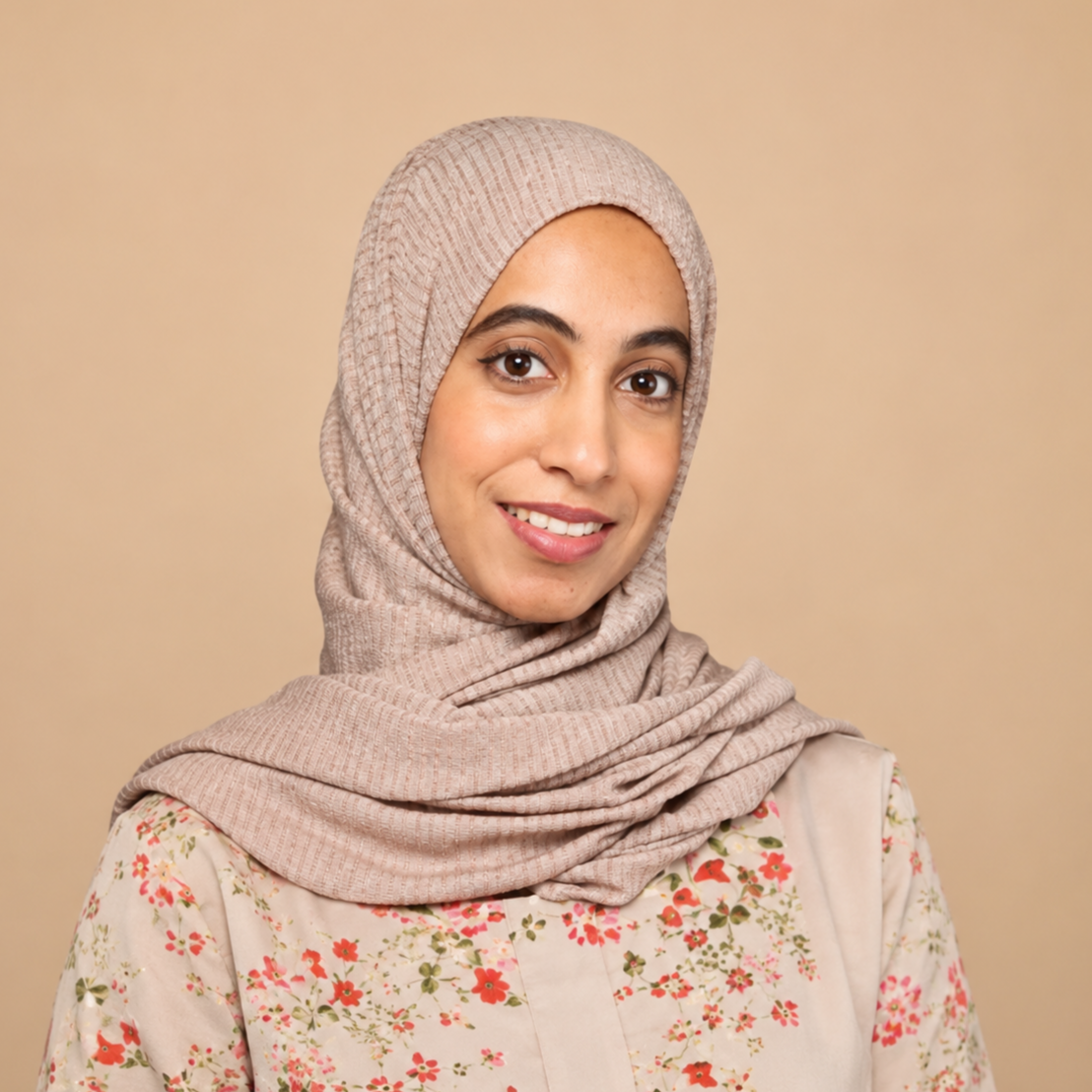 A young woman wearing a beige hijab and a floral blouse, smiling at the camera against a plain beige background.