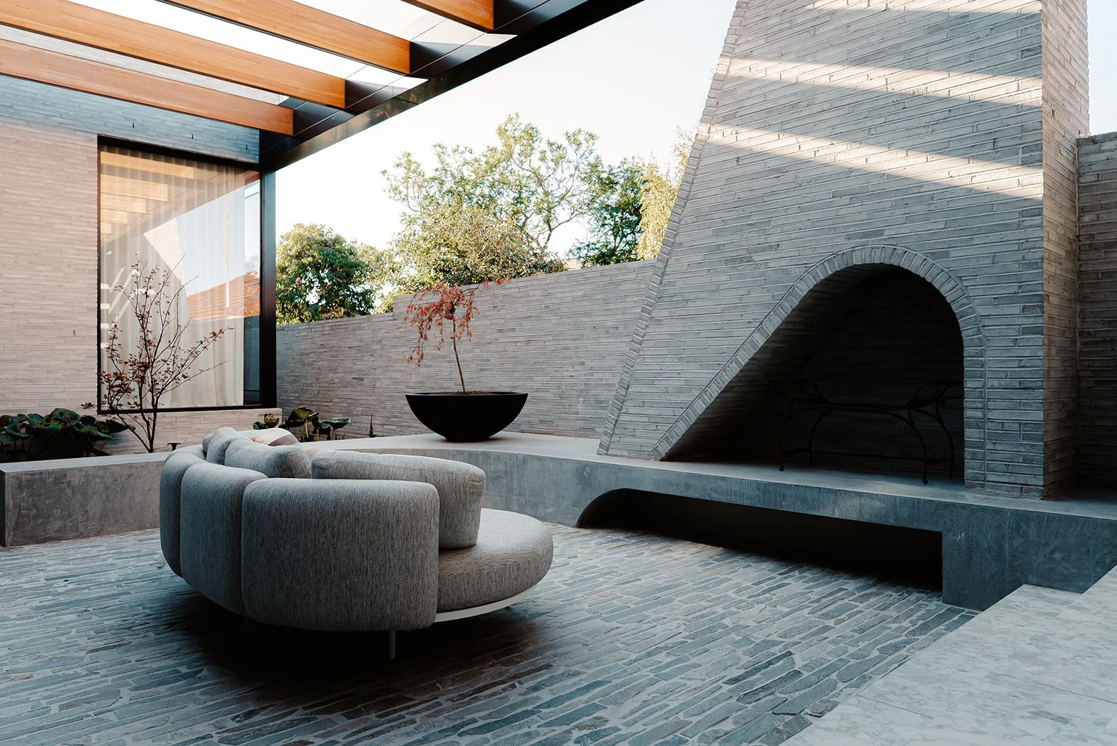 Modern outdoor patio with curved light gray seating, potted plant, brick fireplace, gray brick walls, and trees in the background.
