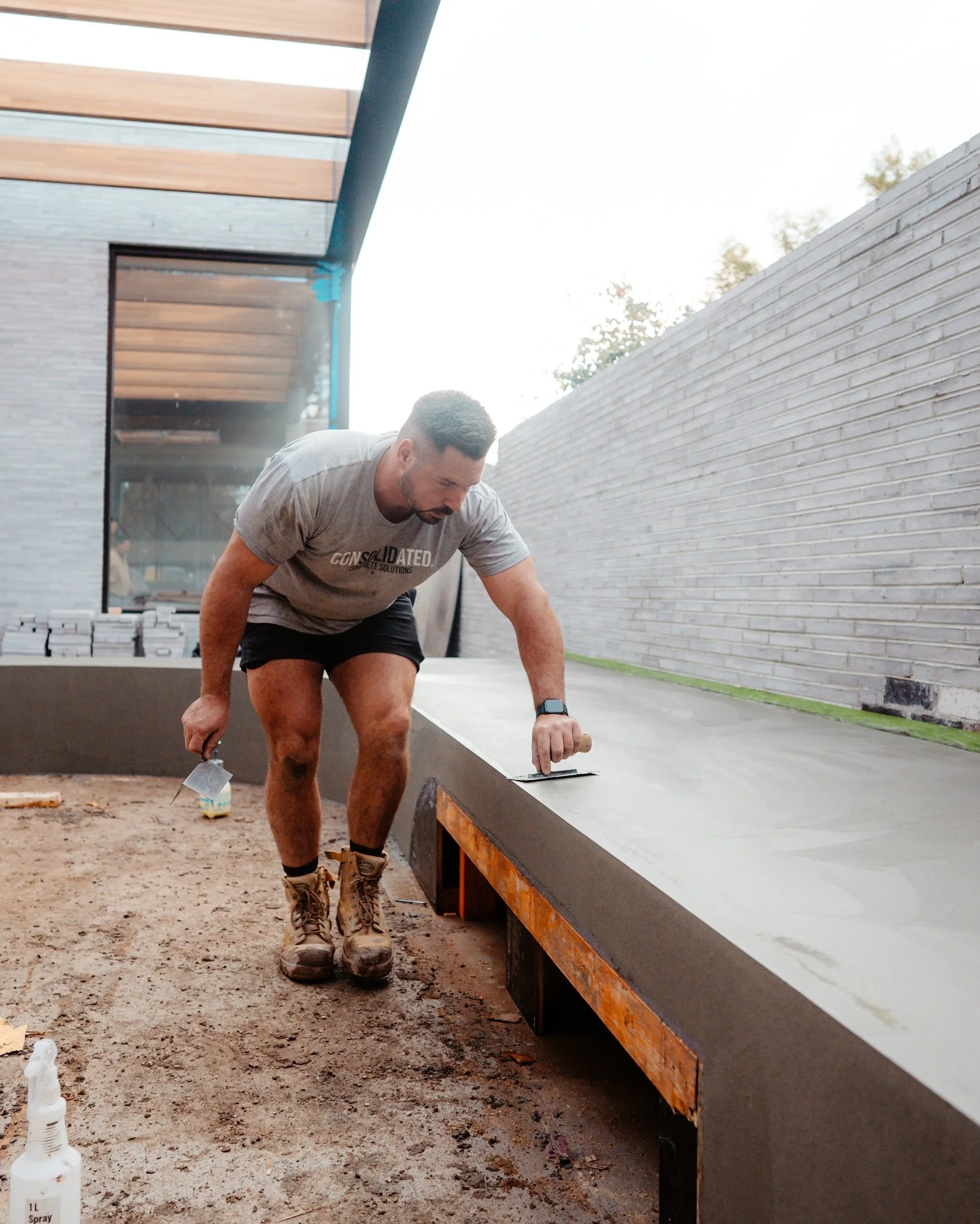 A man working on a construction project outdoors, wearing work boots, a smartwatch, and a t-shirt that says 'CONSOLIDATED,' bent over measuring or inspecting a large flat concrete surface against a wooden formwork, with a modern building wall and trees in the background.
