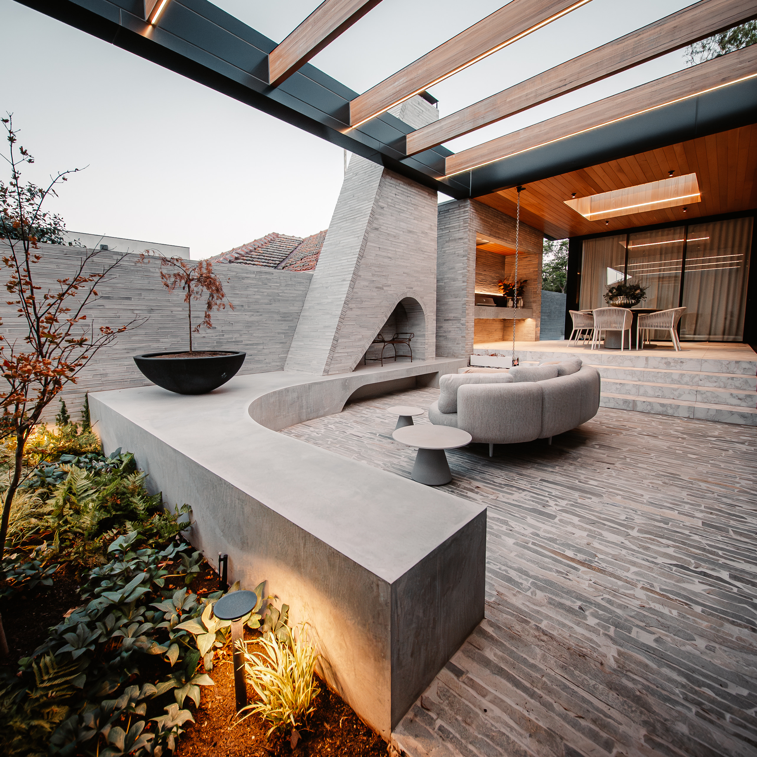 Modern house exterior with a covered patio, wooden beams, brick walls, outdoor furniture, and garden lighting.