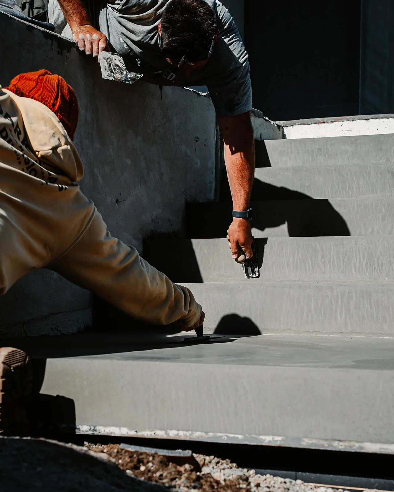 Two workers are applying a smooth layer of concrete to stairs at a construction site. One worker is kneeling and using a trowel, while the other is leaning over the edge, holding a tool. Shadows are cast on the surface being worked on due to bright sunlight.