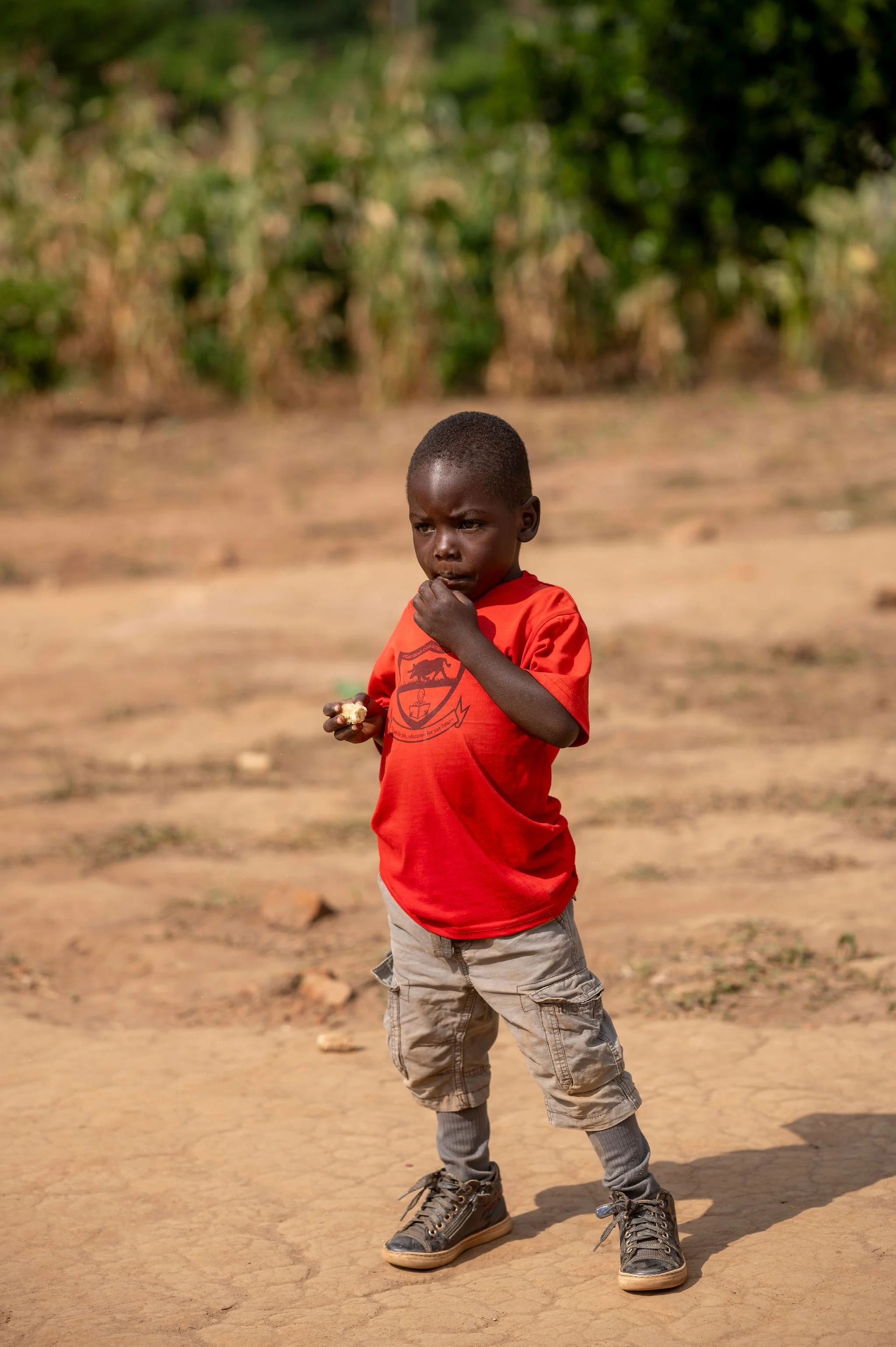 A young boy wearing a red shirt and beige shorts walking on a dusty, cracked ground with a background of green bushes and trees.