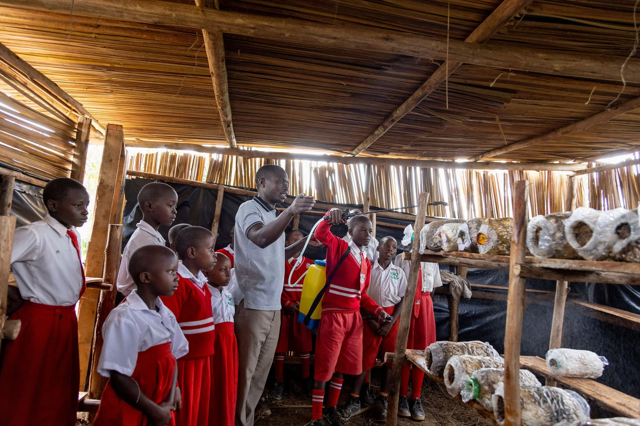 A group of children and an adult inside a wooden structure with a thatched roof, with rocks and bottles on shelves.
