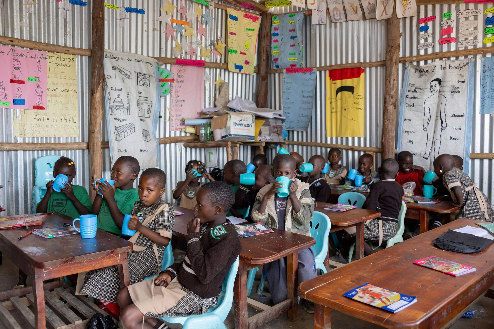A classroom filled with young children sitting at wooden tables, holding blue cups, with educational posters and drawings on the corrugated metal walls behind them.