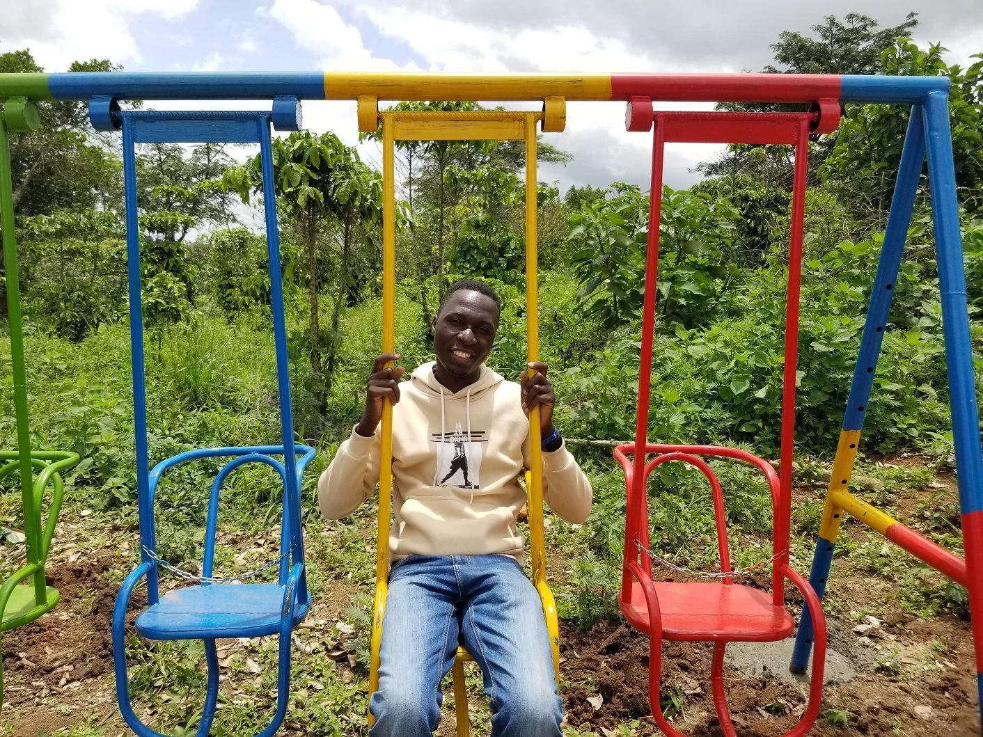 A smiling man sitting on a yellow swing in a colorful playground with green foliage in the background.