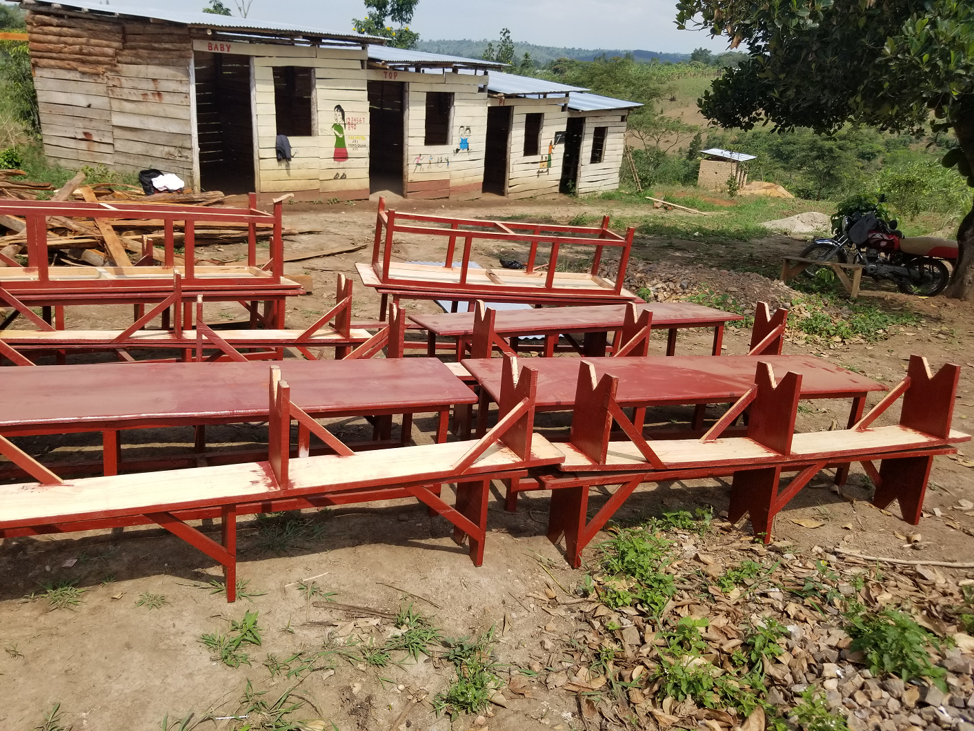 Multiple tables and chairs placed outdoors on dirt ground, with a rustic wooden building featuring painted child-themed artwork in the background, and a motorcycle parked near a tree on the right side.