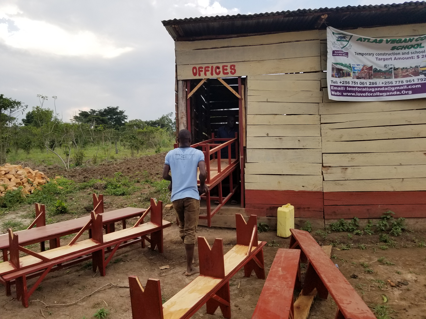 A man walking up a small ramp into a wooden building labeled 'OFFICES' with benches in the outdoor area. The building has a sign indicating it is part of Atlas Vegan Community School, under construction, with contact information visible.