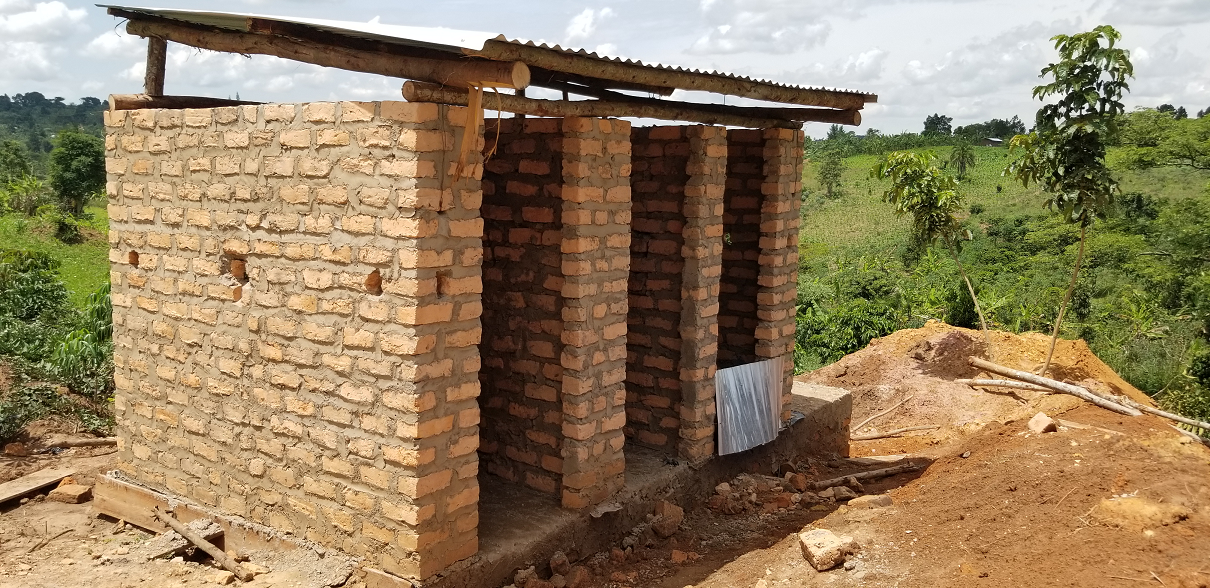 A small brick structure under construction in a rural area with green trees and hills in the background.