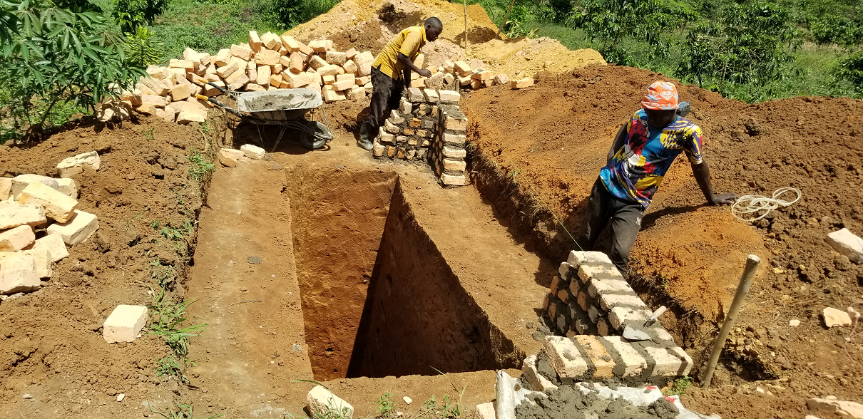 Two workers building a brick wall in a deep excavation site outdoors, with piles of dirt and additional bricks around, and green foliage in the background.