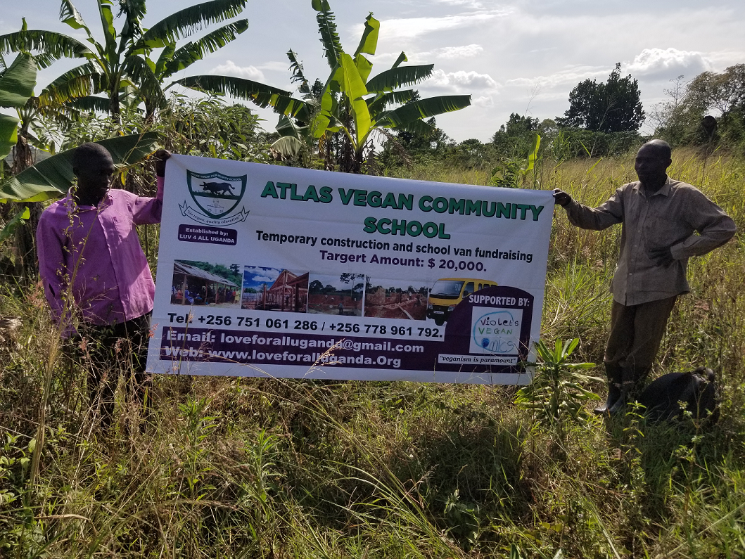 Two men standing outdoors in a grassy area with banana trees, holding a banner for the Atlas Vegan Community School, which mentions its temporary construction and fundraising goals, along with contact information and supporting organizations.