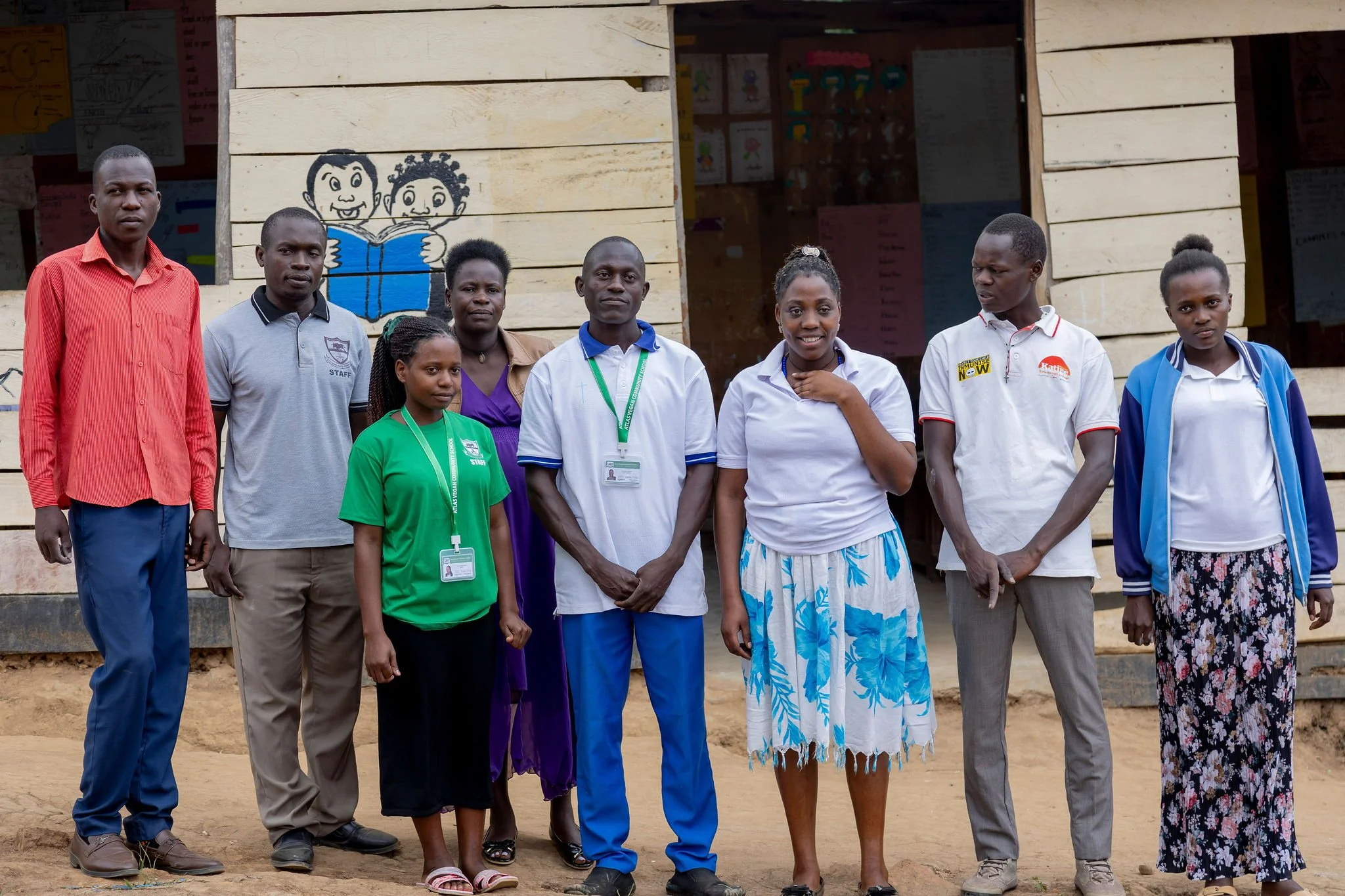 Group of nine people standing in front of a wooden building with colorful drawings of children reading painted on it.