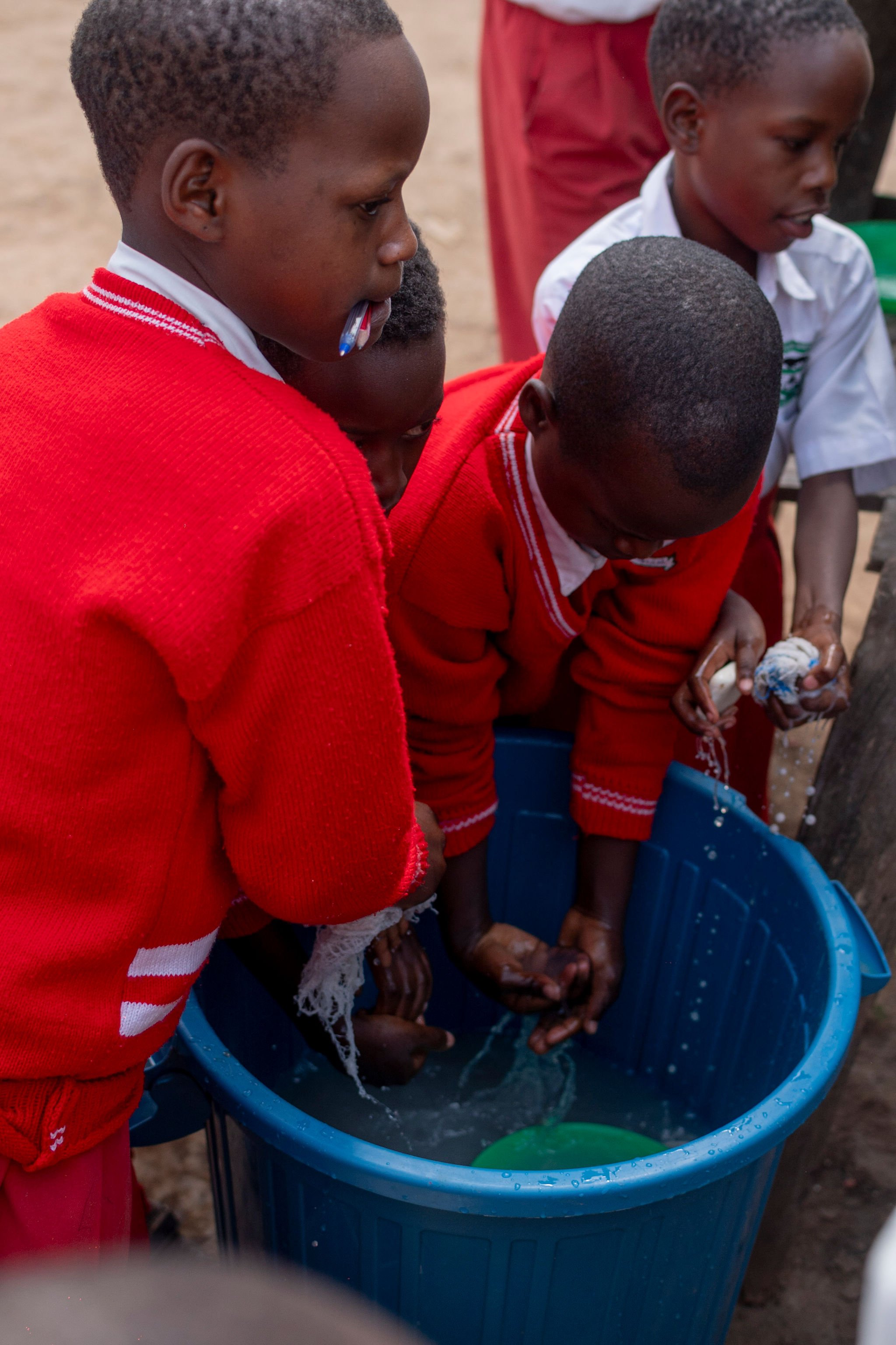 Children with dark skin washing their feet in a blue plastic basin outdoors, some wearing red sweaters and white shirts.