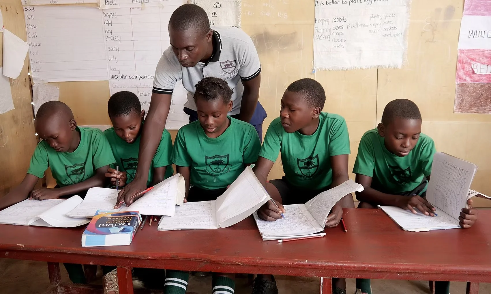 Five boys in green school uniforms sitting at a wooden desk, studying from open notebooks in a classroom, with a teacher assisting them.