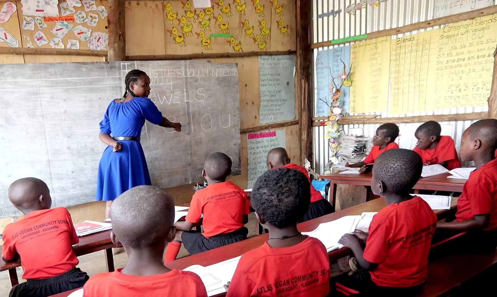 A classroom scene with a female teacher in a blue dress instructing students, all wearing red uniforms, sitting at desks with open books. The classroom walls are decorated with posters, drawings, and handwritten notes.