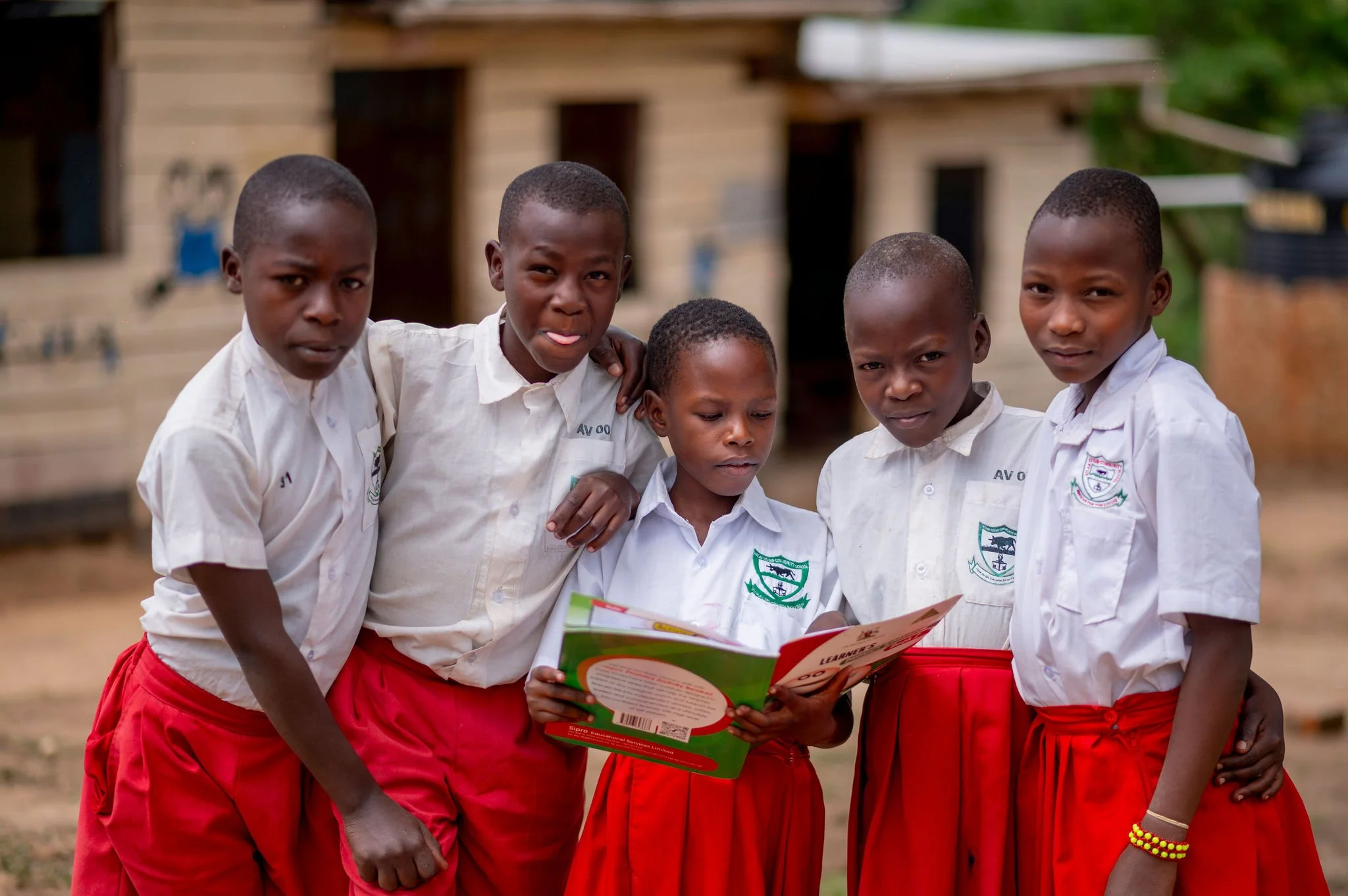 Five schoolchildren in uniform standing outdoors, looking at a book together, with a wooden building in the background.