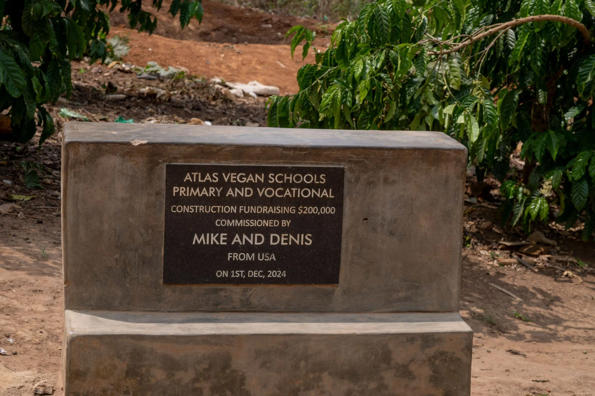 A stone plaque in an outdoor setting with green foliage in the background. The plaque is inscribed with information about Atlas Vegan Schools, including details about primary and vocational education, a construction fundraising goal of $200,000, and 