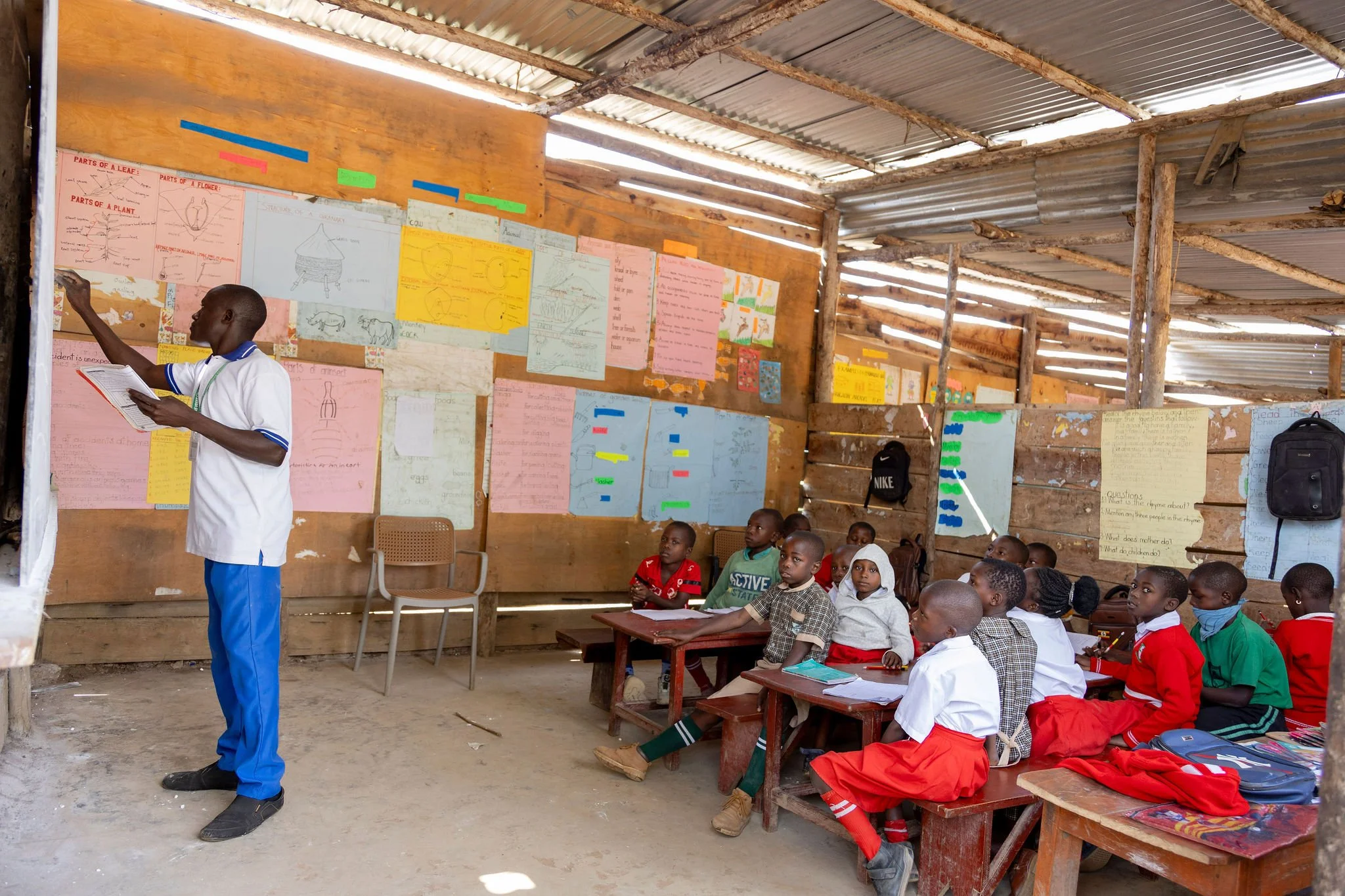 A classroom of young students sitting on wooden benches, listening to a teacher pointing at a board in a school built with wooden planks and a metal roof.
