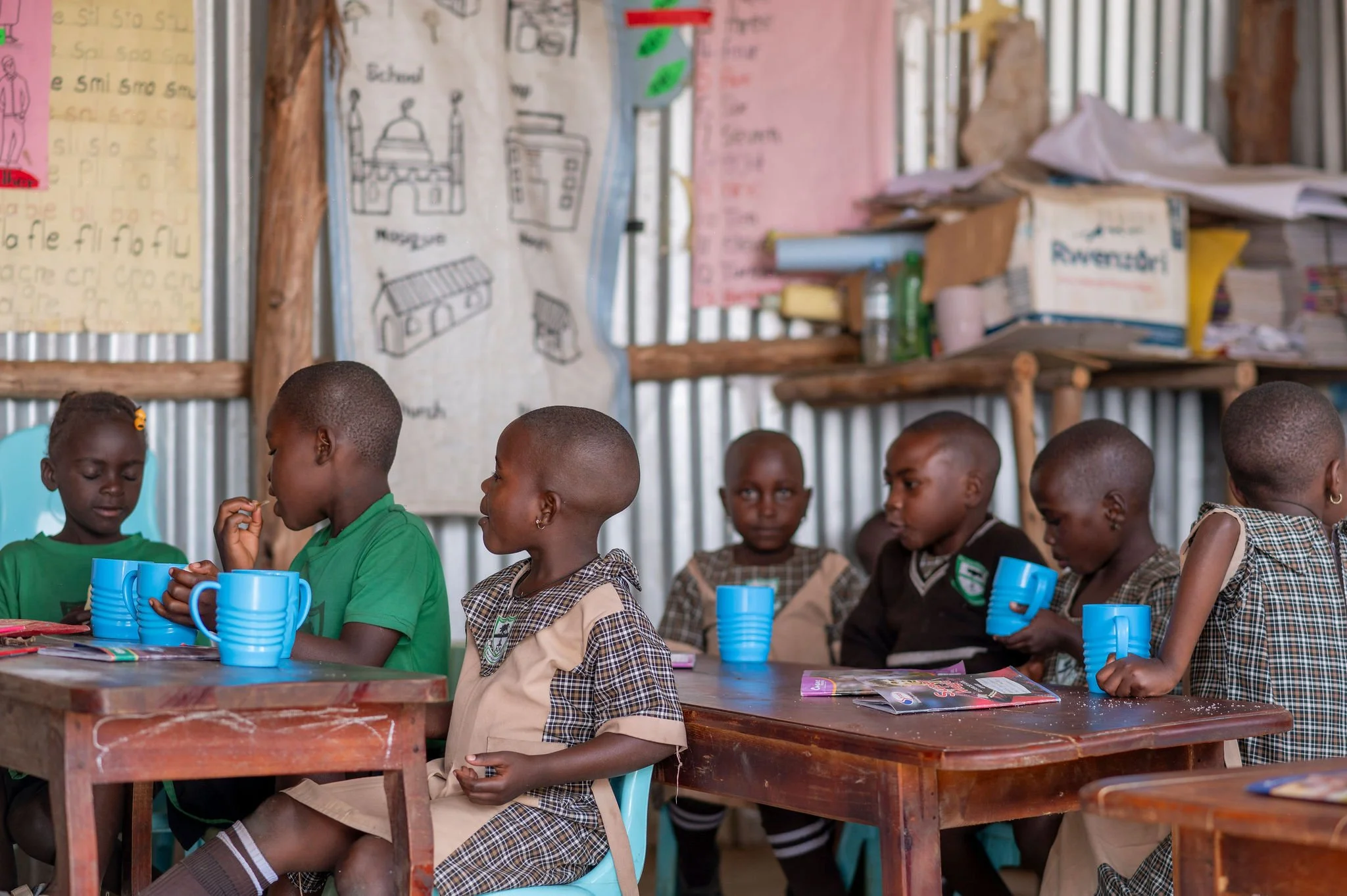Group of children sitting at a wooden table with blue cups, in a classroom with posters and drawings on the walls.
