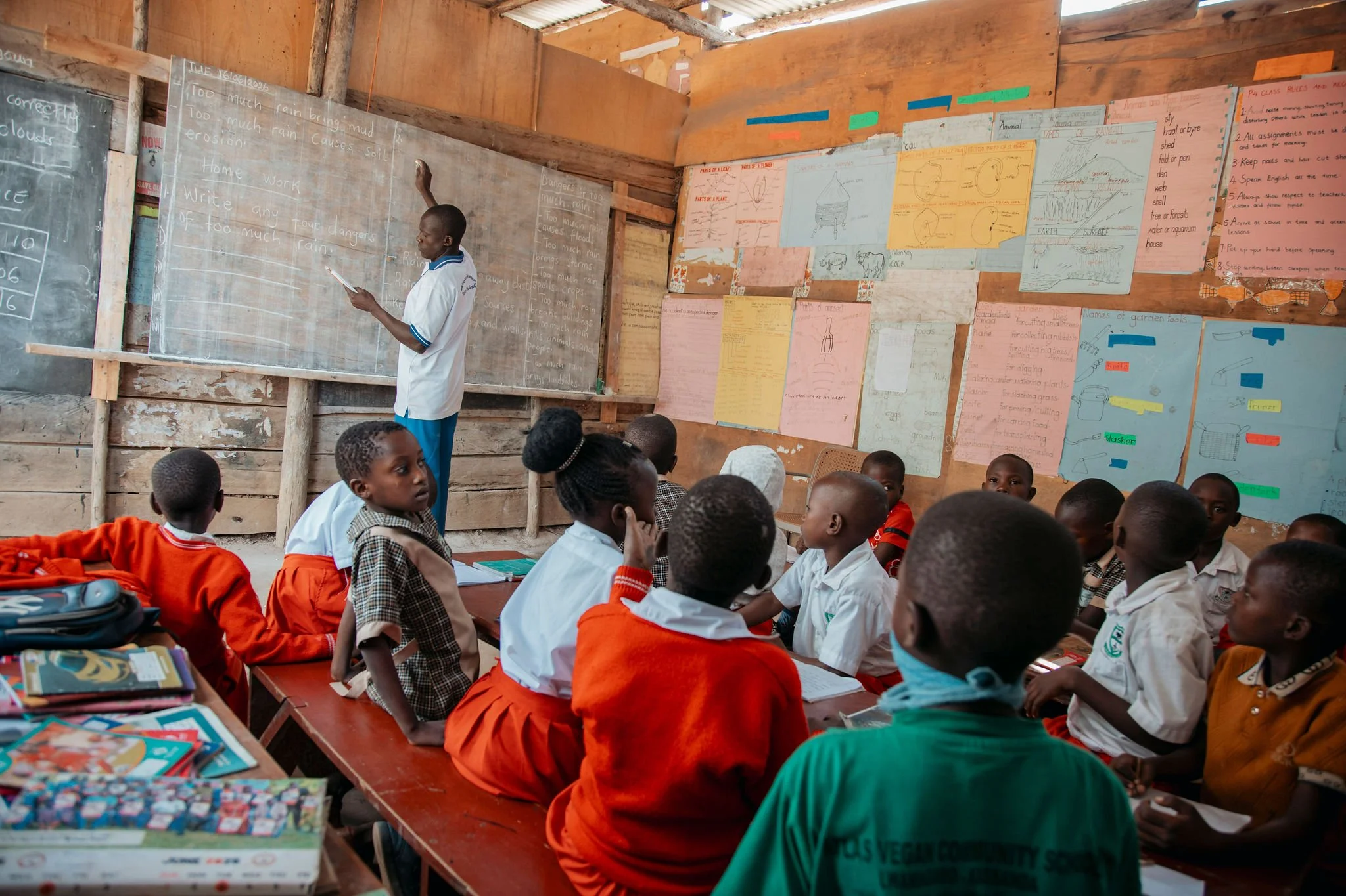 A classroom filled with young students seated at desks, listening to a teacher who is standing by a chalkboard with writing on it. The classroom has colorful posters on the walls and a wooden ceiling.