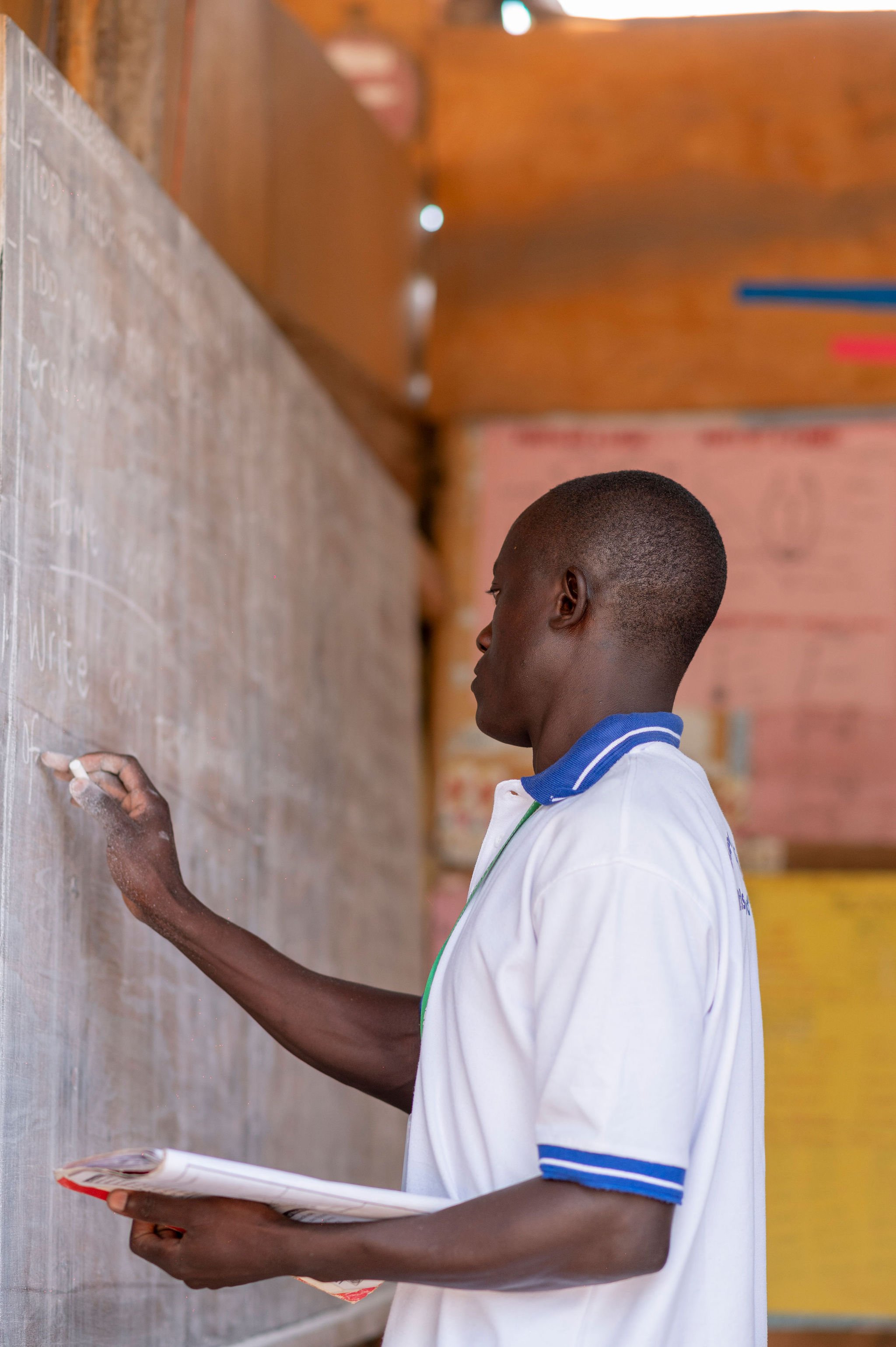 A young boy in a white shirt with blue trim writing or drawing on a chalkboard or whiteboard in a classroom.