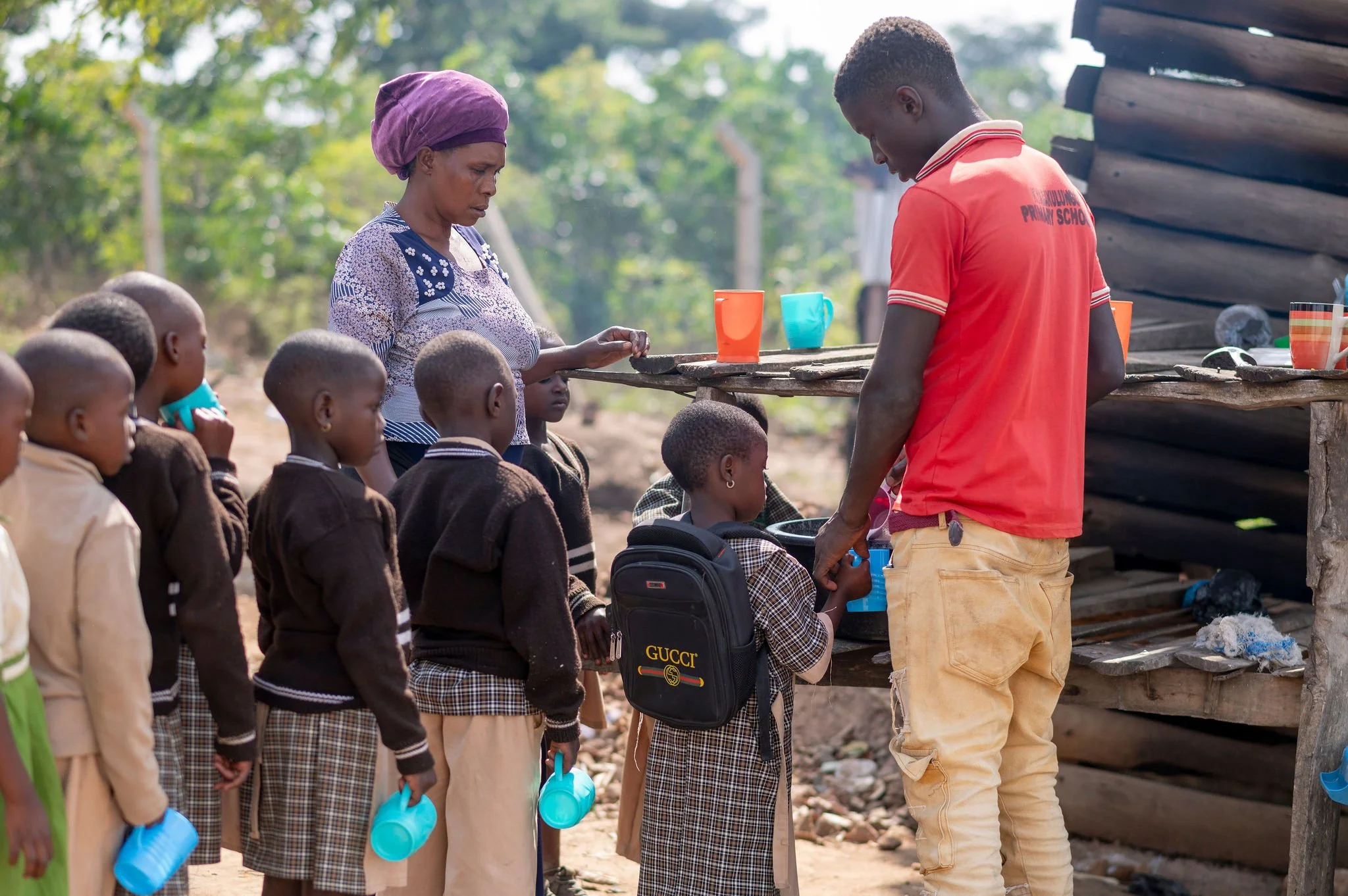 A woman and a man serving food to children in an outdoor setting. The children are in line, holding blue water bottles and wearing school uniforms. The woman wears a patterned dress and purple headscarf, while the man wears a red polo shirt and beige