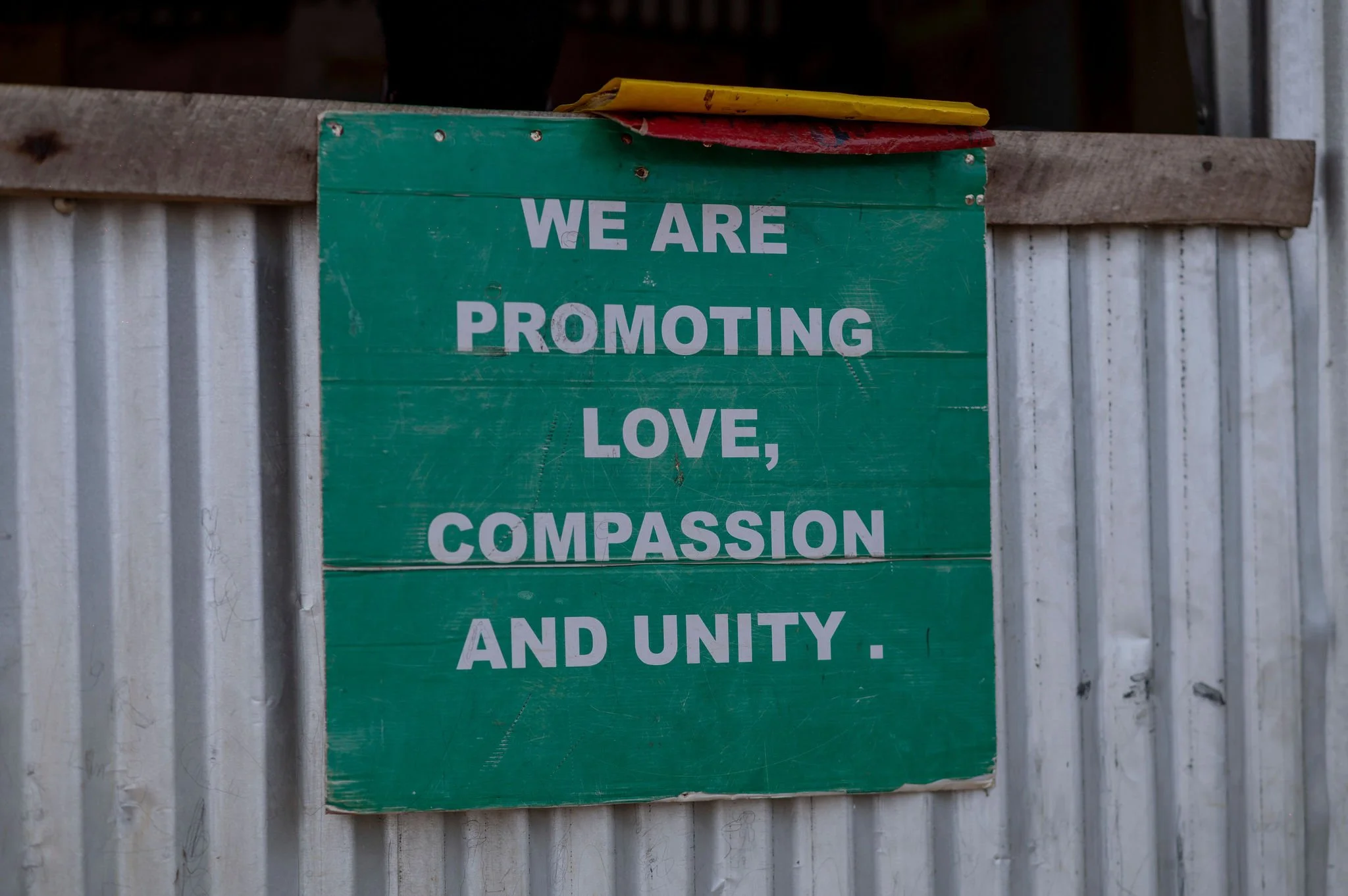 A green wooden sign with white text that reads: "WE ARE PROMOTING LOVE, COMPASSION AND UNITY." The sign is mounted on a corrugated metal surface, and there are some yellow and red objects placed on top of it.