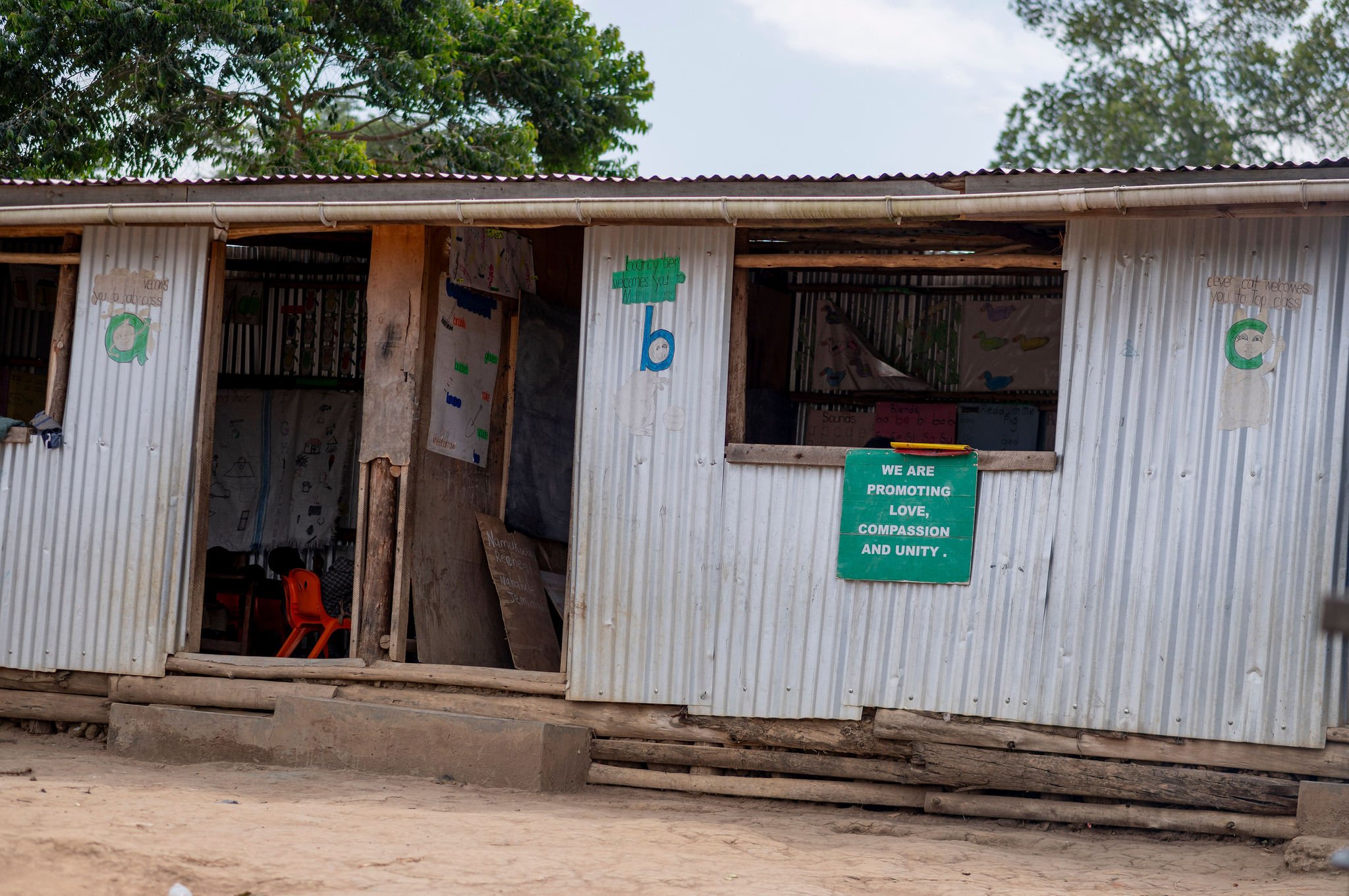 A small building made of corrugated metal sheets with a doorframe and a window, located in a rural area with dirt ground and green trees in the background. Several signs are attached to the building, including a green sign that reads "We are promotin