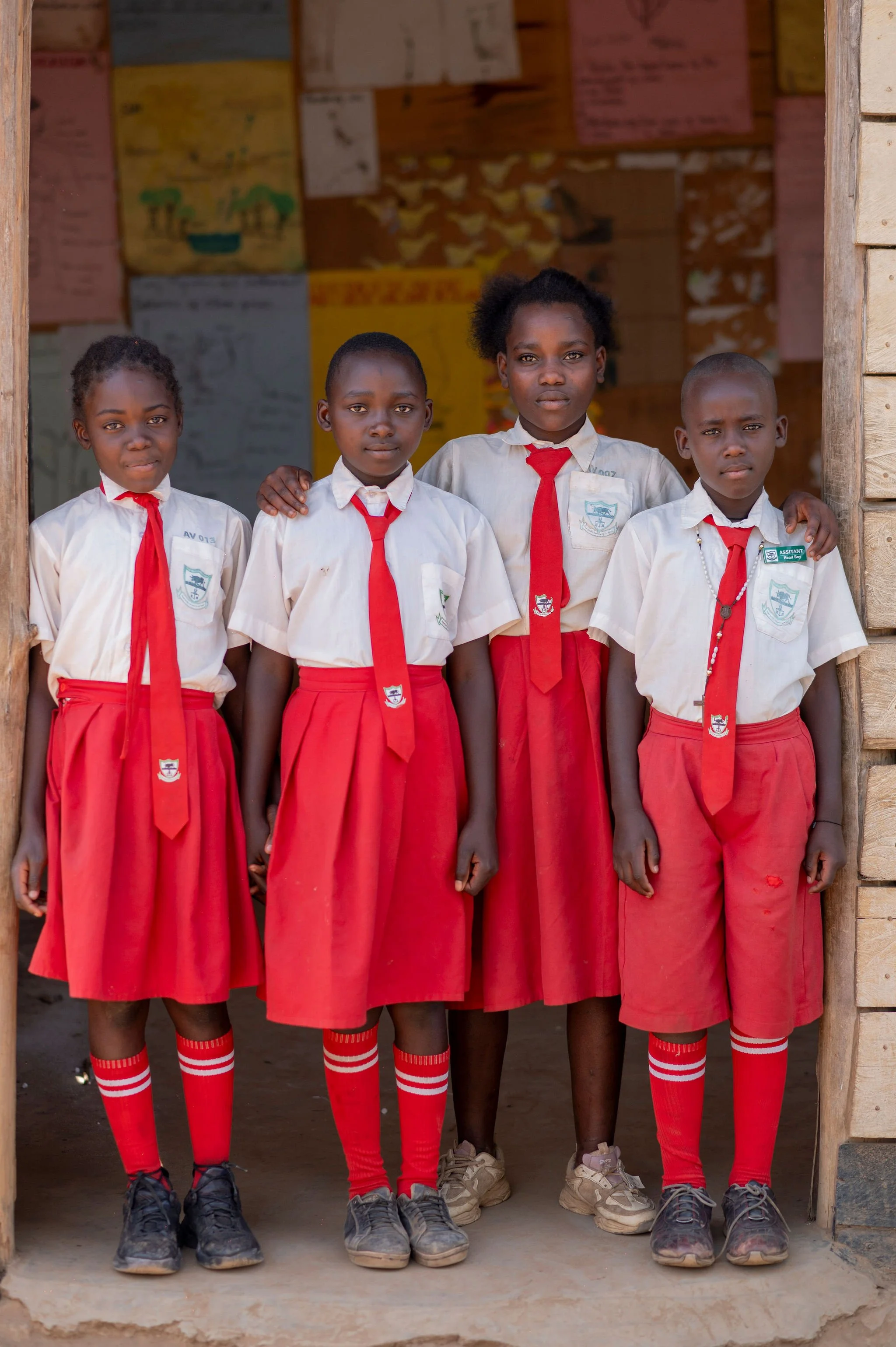 Four young schoolgirls in uniform standing in front of a classroom door with their arms around each other. They are wearing white shirts, red skirts, and red and white striped knee-high socks, with some girls also wearing red ties. The background sho