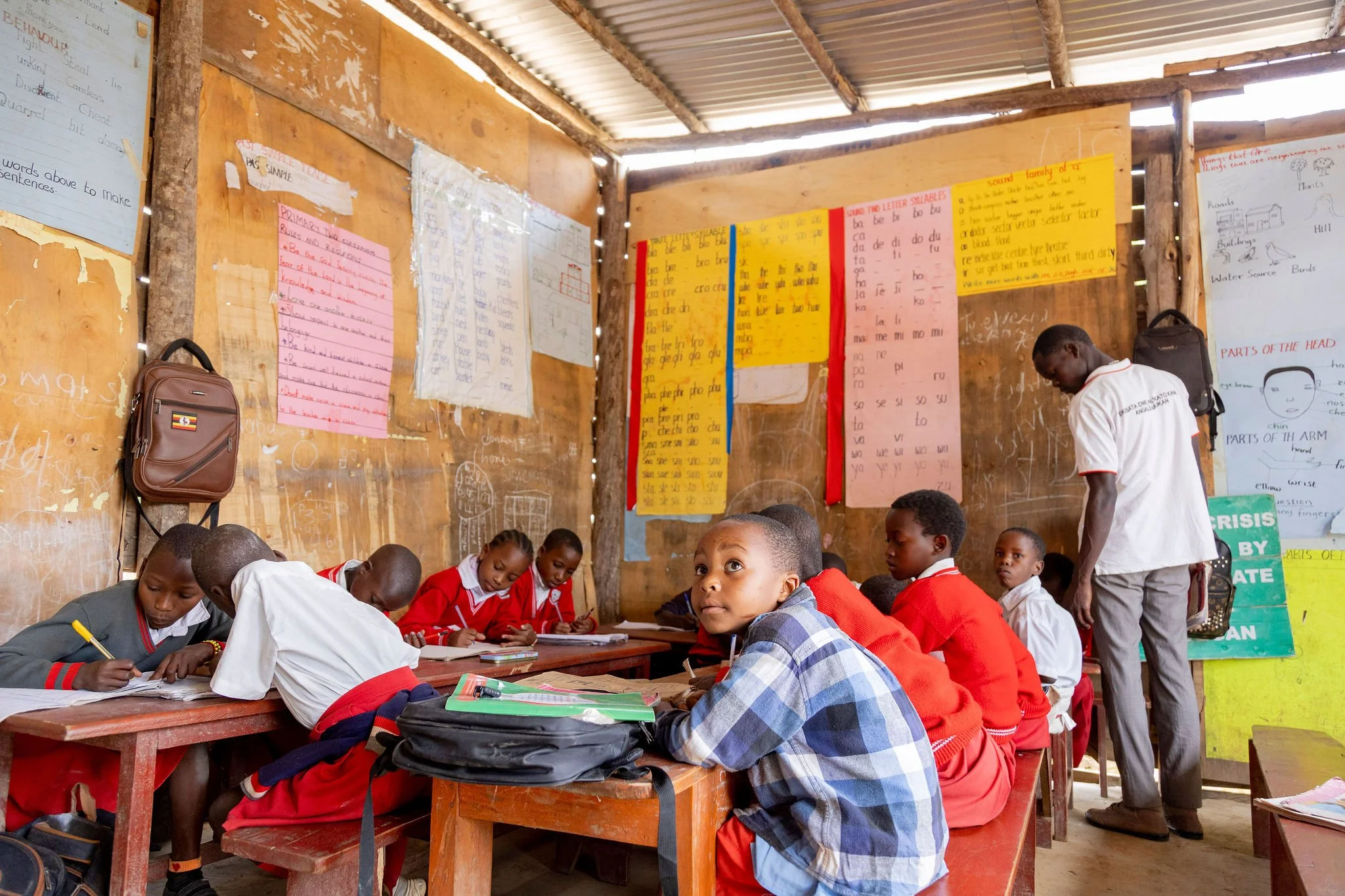 A classroom with students seated at desks, working on assignments, and a teacher standing nearby. The classroom walls display educational posters and charts.
