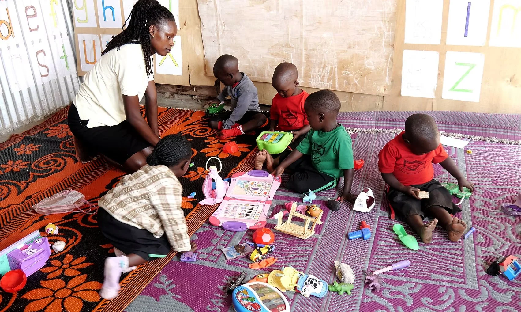 A woman and five children playing with toys and educational materials on the floor of a classroom with colorful alphabet drawings on the wall.