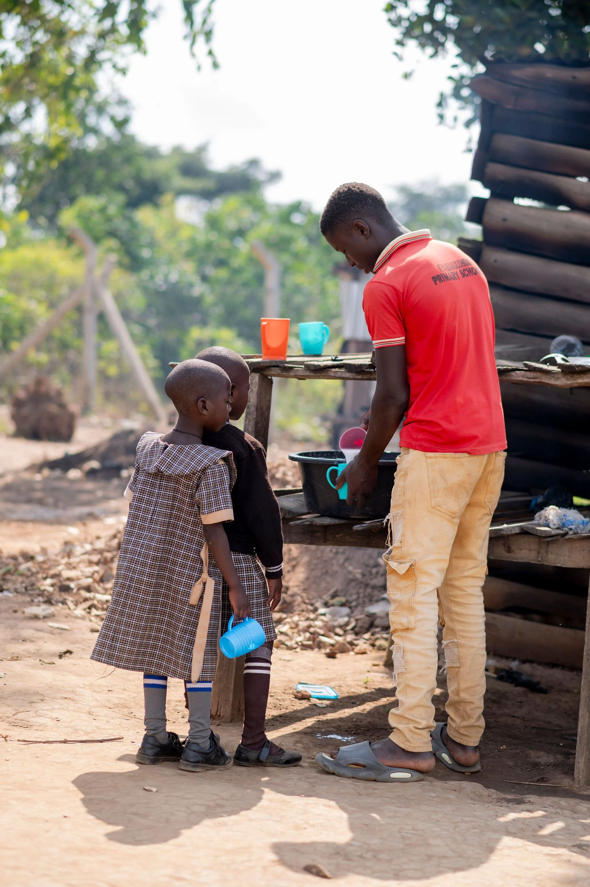 A man and two children standing outdoors with one child holding a blue watering can, near a wooden structure, with trees and a bright sky in the background.