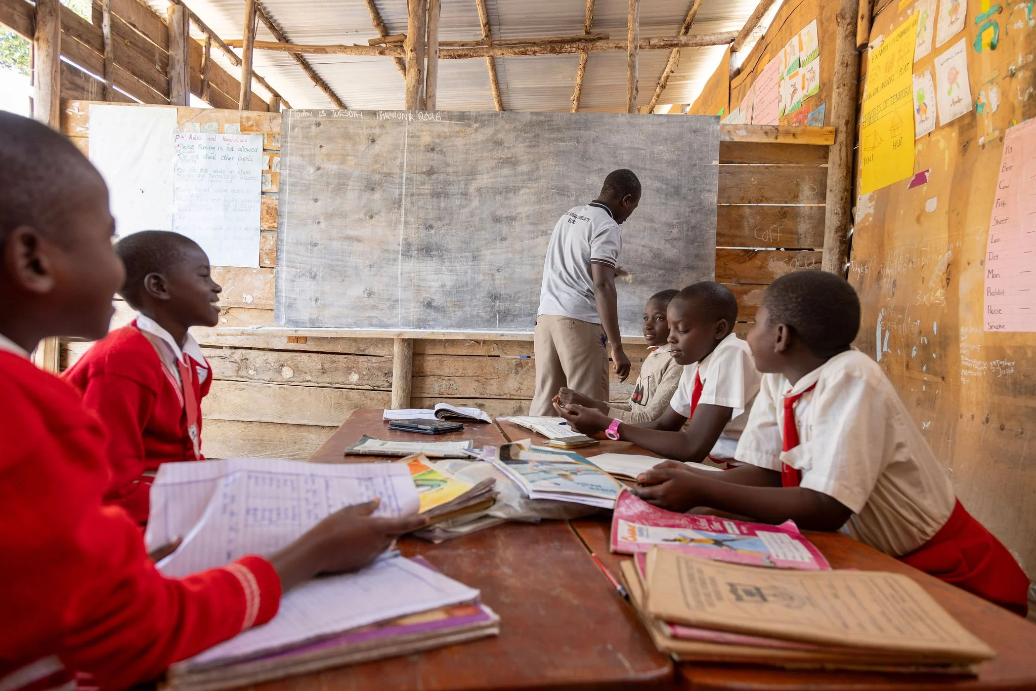A classroom scene with students sitting at a table, engaging with books and papers, while a teacher writes on the blackboard. The classroom has unfinished wooden walls and colorful educational posters.