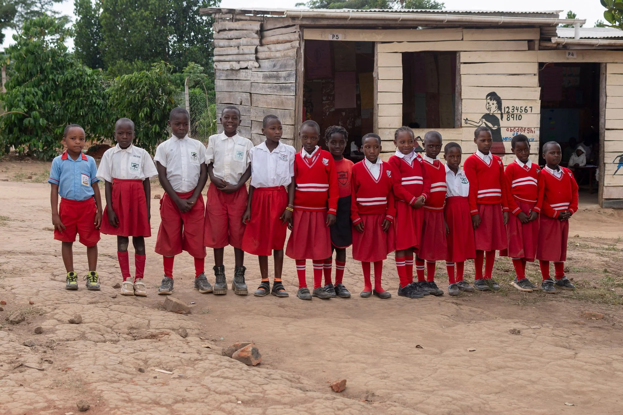 A group of school children standing in a line outdoors in front of a wooden school building. The children are wearing school uniforms, some in white shirts and others in red sweaters or red skirts. They are facing the camera, and there are trees and 